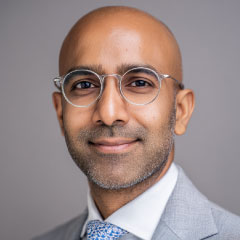 Portrait of an Indian man in glasses and a grey suit, smiling slightly.