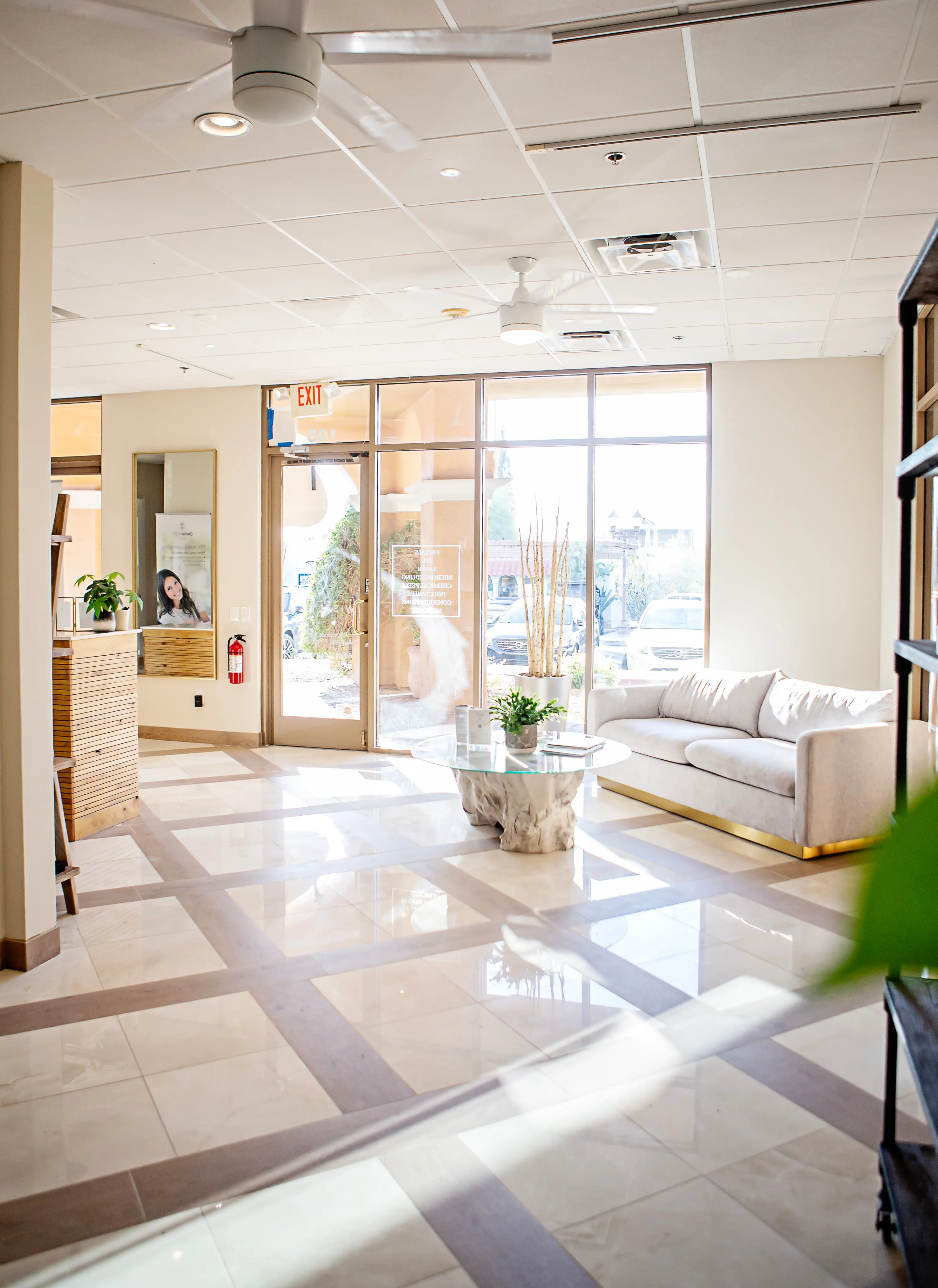 Bright and modern medical clinic waiting area with large windows, a neutral-toned couch, a decorative glass table, and indoor plants.