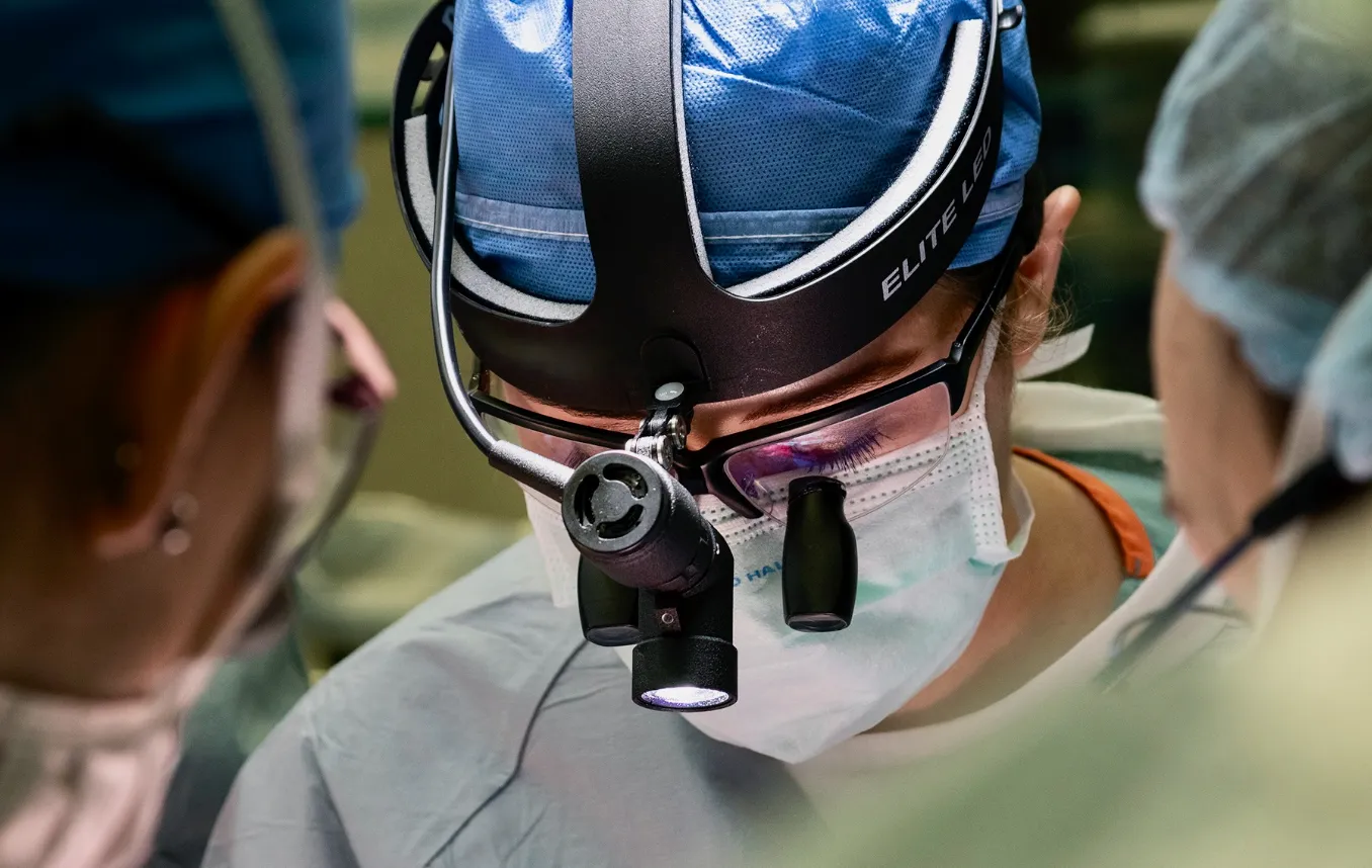 Surgeon wearing headlamp and magnifying glasses focuses during an operation, surrounded by medical staff.