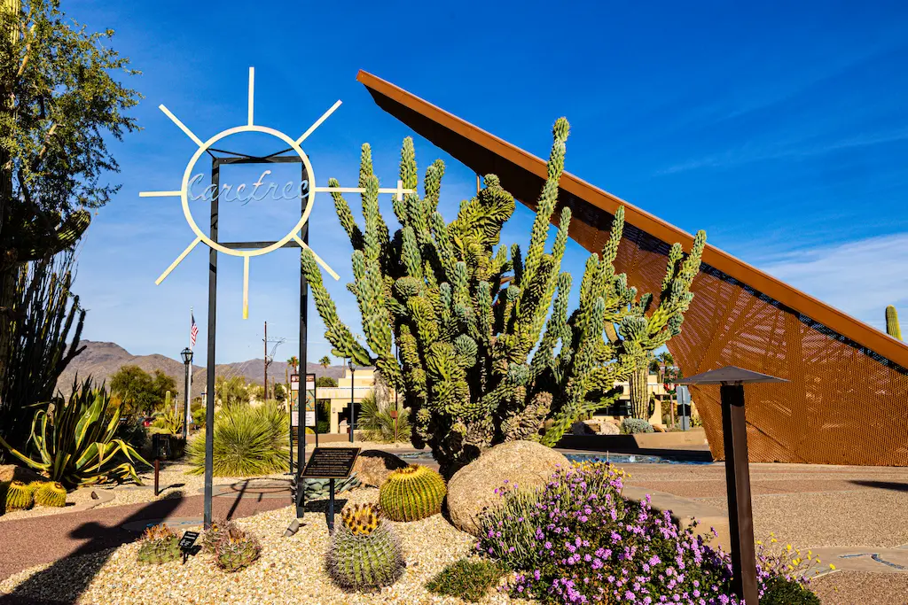 Desert landscape with a large cactus and a sun sculpture displaying "Carefree" set against a clear blue sky. Mountains and buildings in the background.