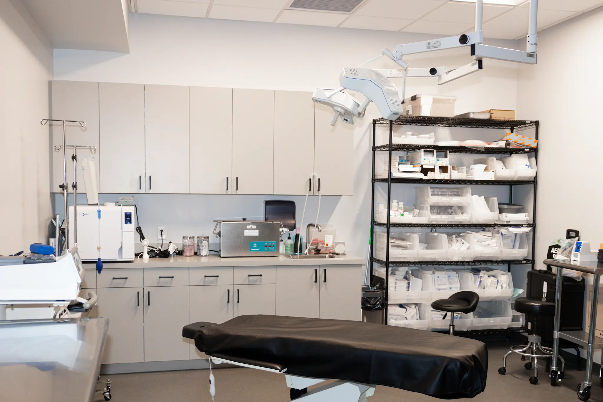 A clean, organized medical examination room. Cabinets are filled with equipment. A black examination table and shelves with supplies are visible.