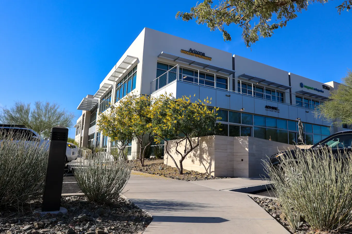 Modern white office building with large windows against a clear blue sky. Desert landscaping with small trees and bushes line the pathway.