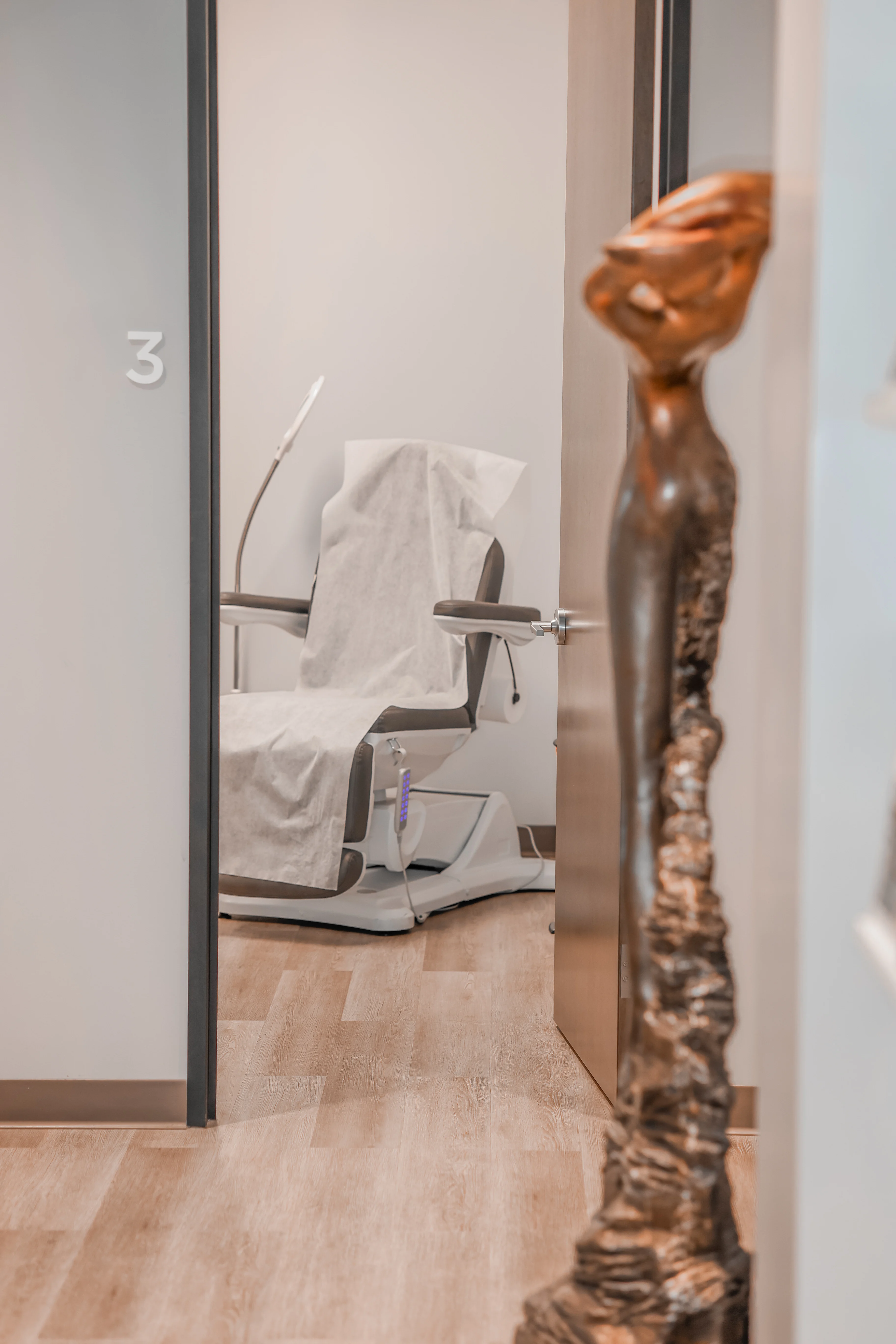 View through doorway to treatment room with modern exam chair at Metropolitan Plastic Surgery, Scottsdale location, with warm wood floors and sculptural decor.