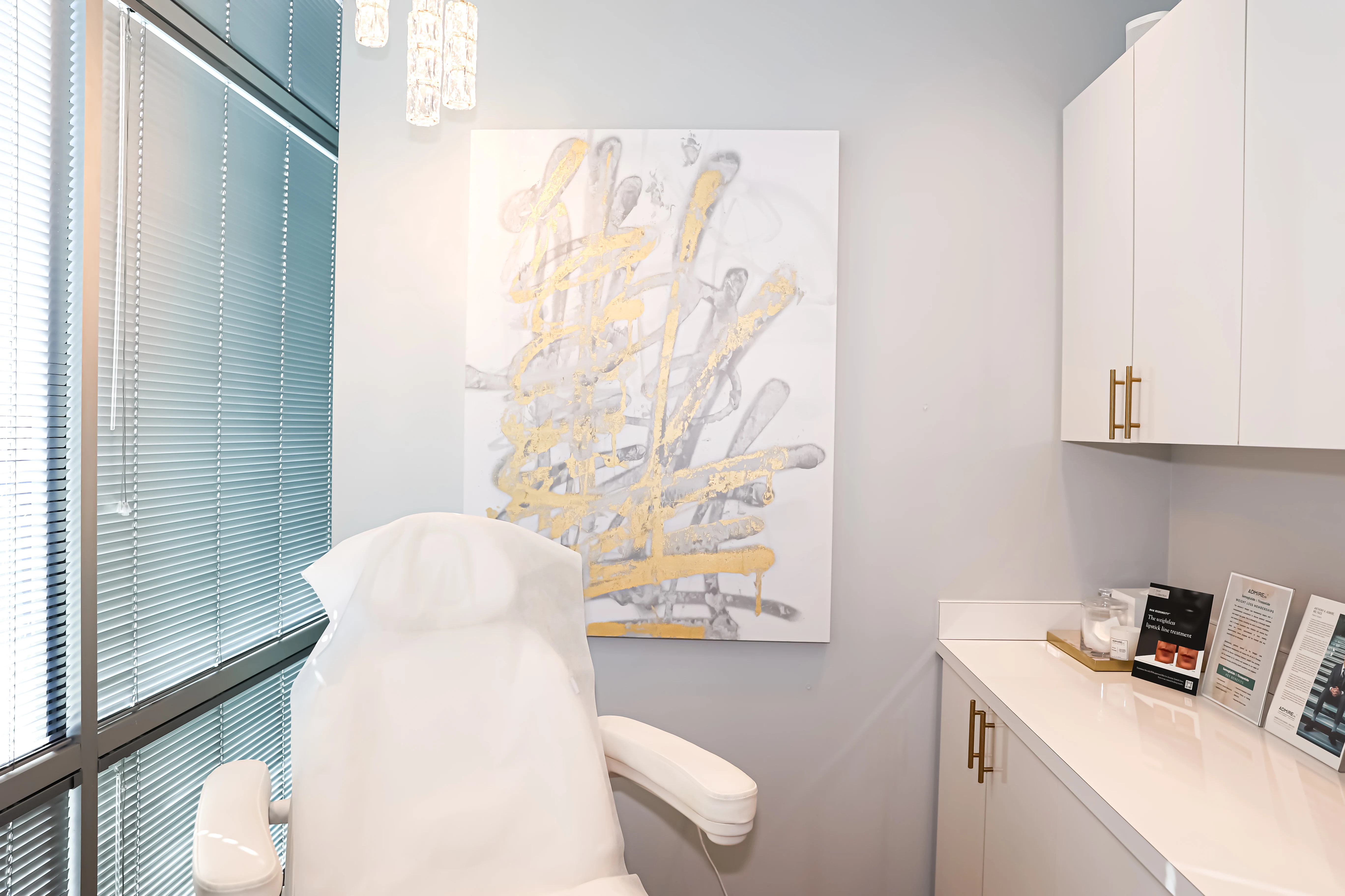 Bright modern Scottsdale treatment room at Metropolitan Plastic Surgery with reclining medical chair, abstract gold and silver art, and sleek cabinetry