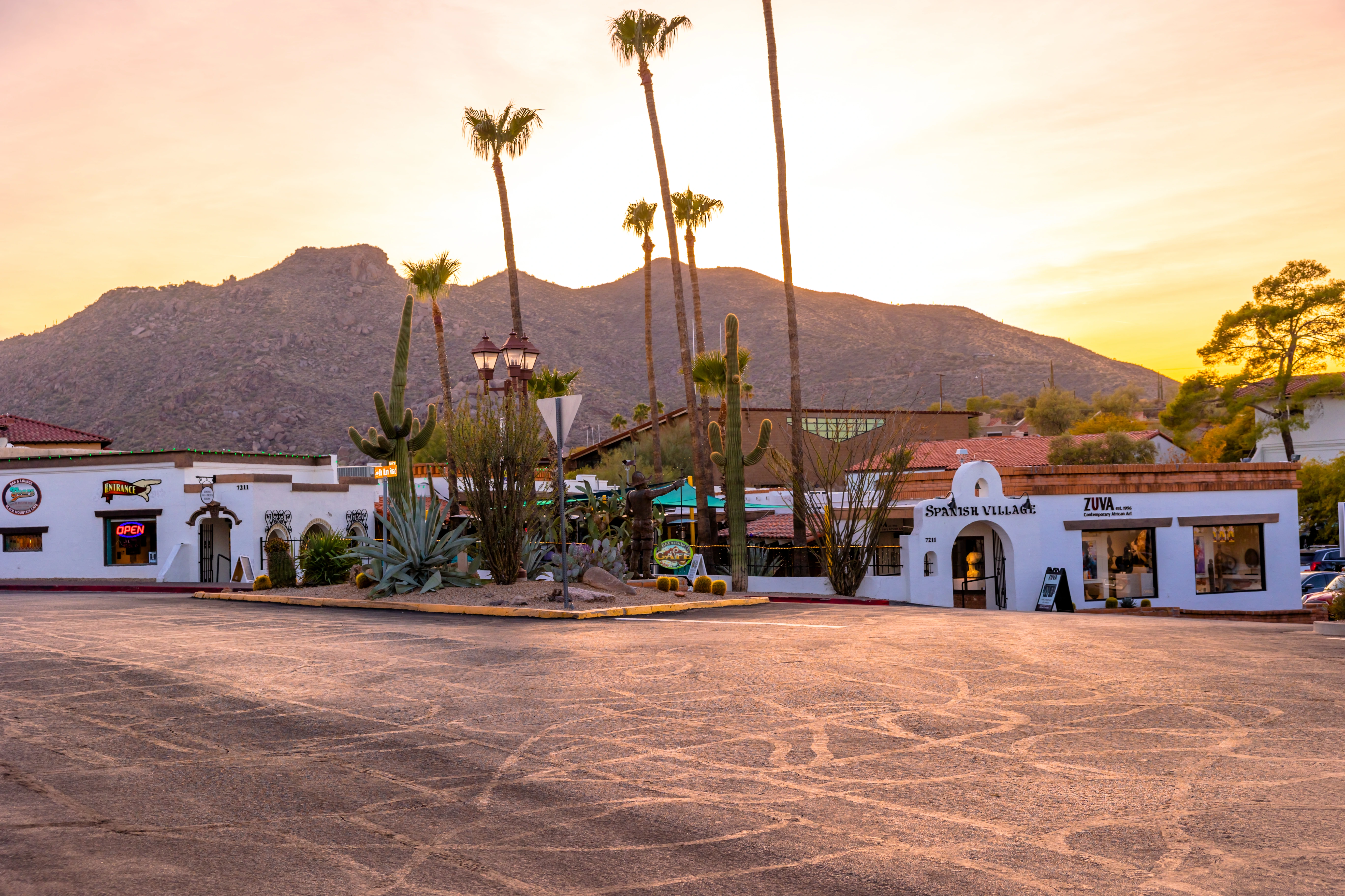 Sunset view of Spanish Village shops in Carefree, Arizona, with tall palm trees, desert landscaping, and Black Mountain in the background.