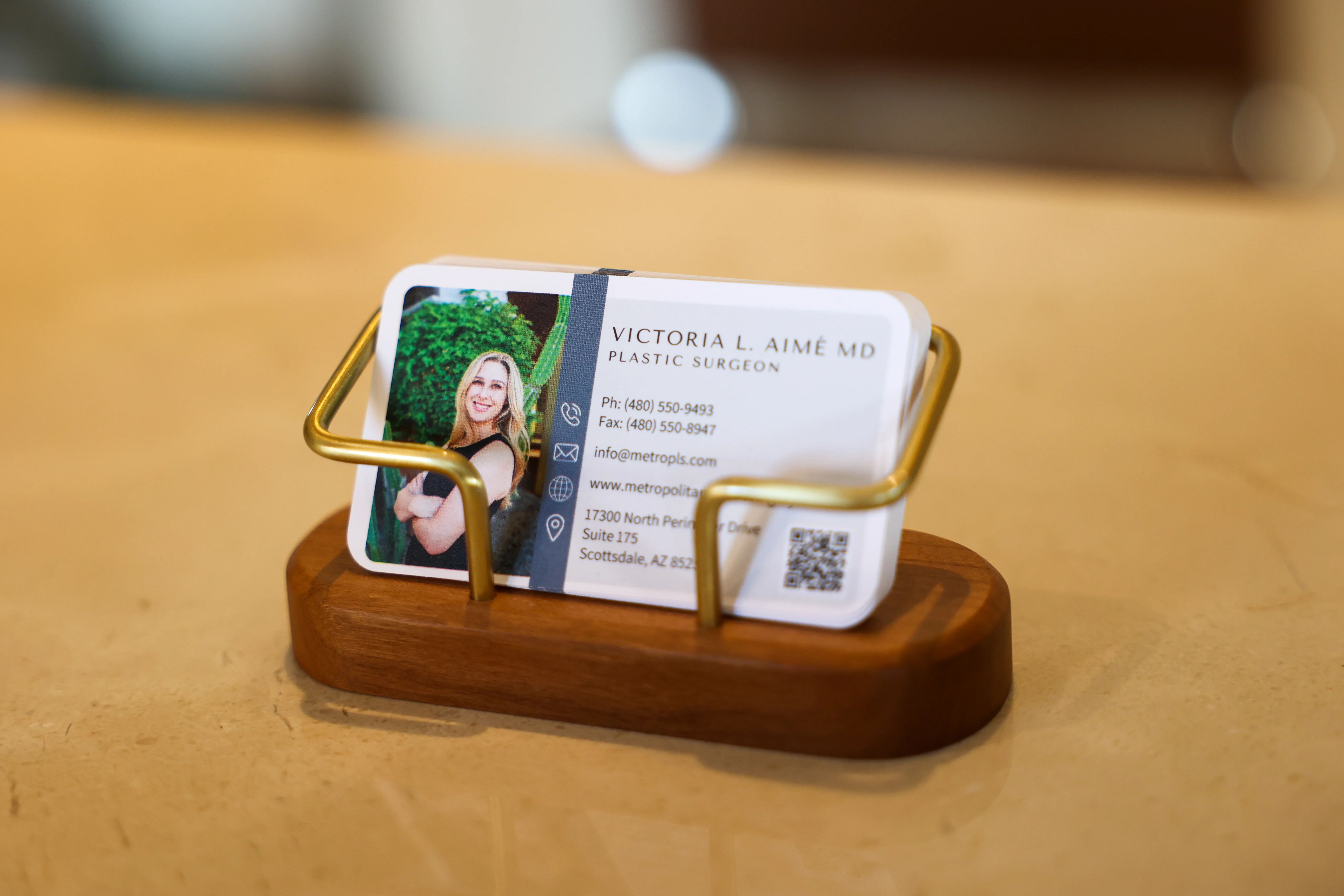 Close-up of a Metropolitan Plastic Surgery business card for Dr. Victoria Aimé, displayed in a wooden and brass holder on the office reception desk.
