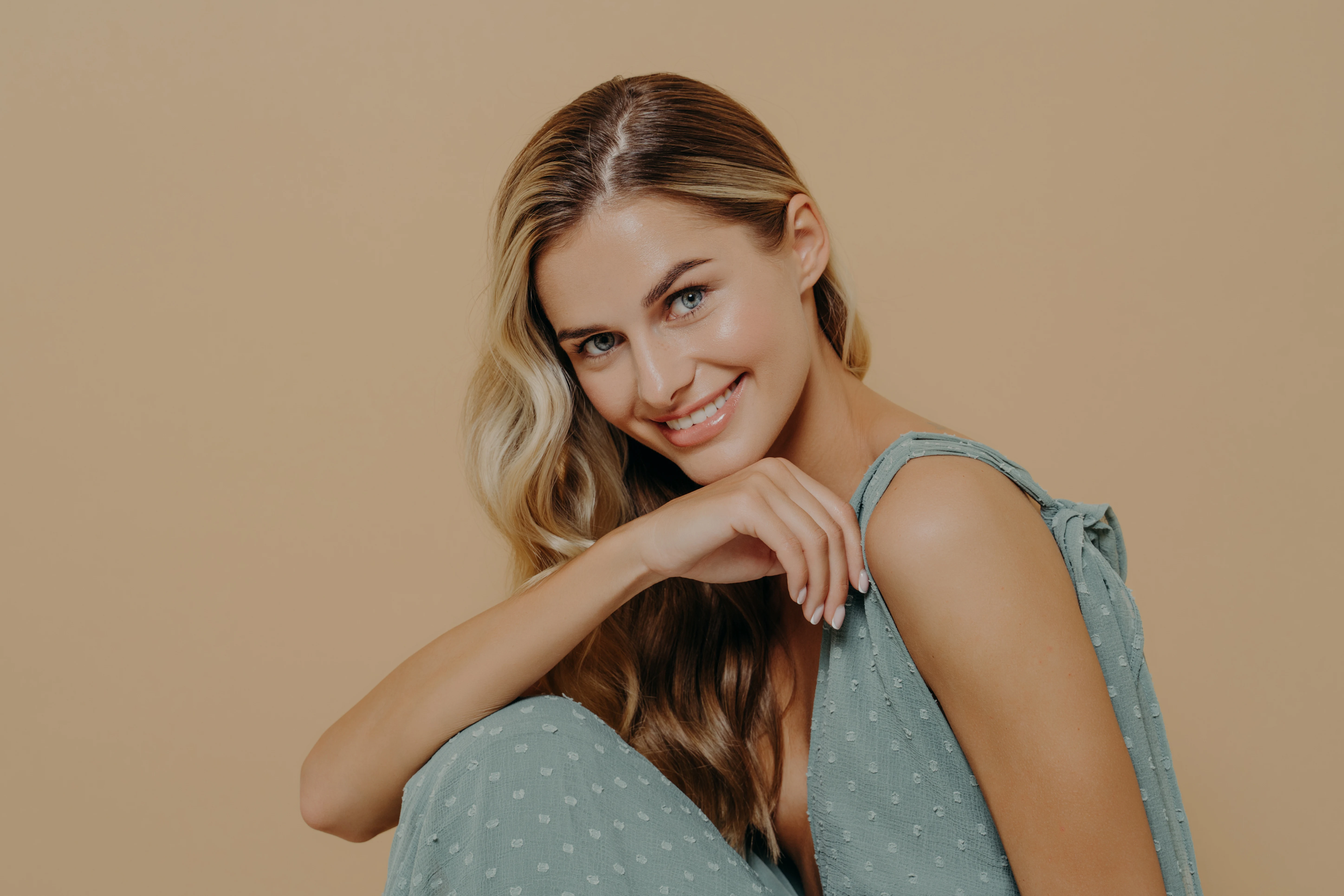 Woman smiling confidently with even-toned, glowing skin after microneedling, seated in a light teal dress against a neutral background.