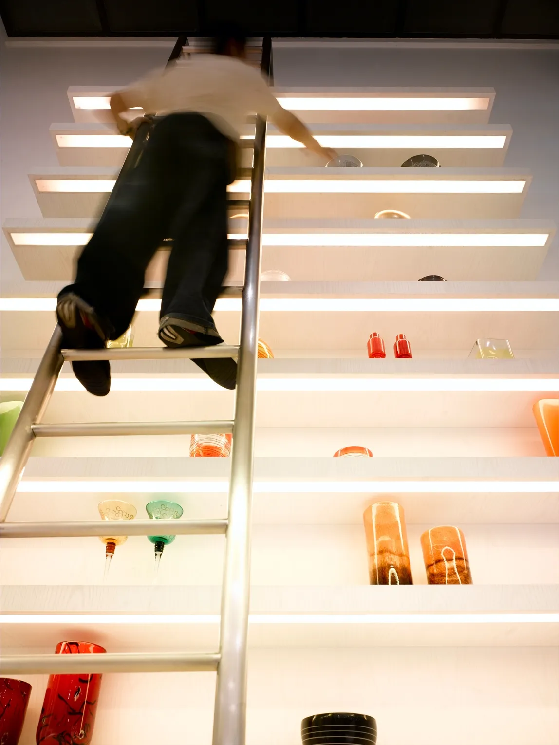 Persona che sale una scala metallica verso scaffali alti con oggetti decorativi colorati.