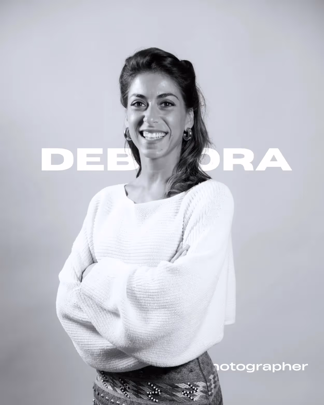 Donna sorridente con maglione bianco e braccia incrociate davanti a sfondo grigio con testo 'DEBORAH'.