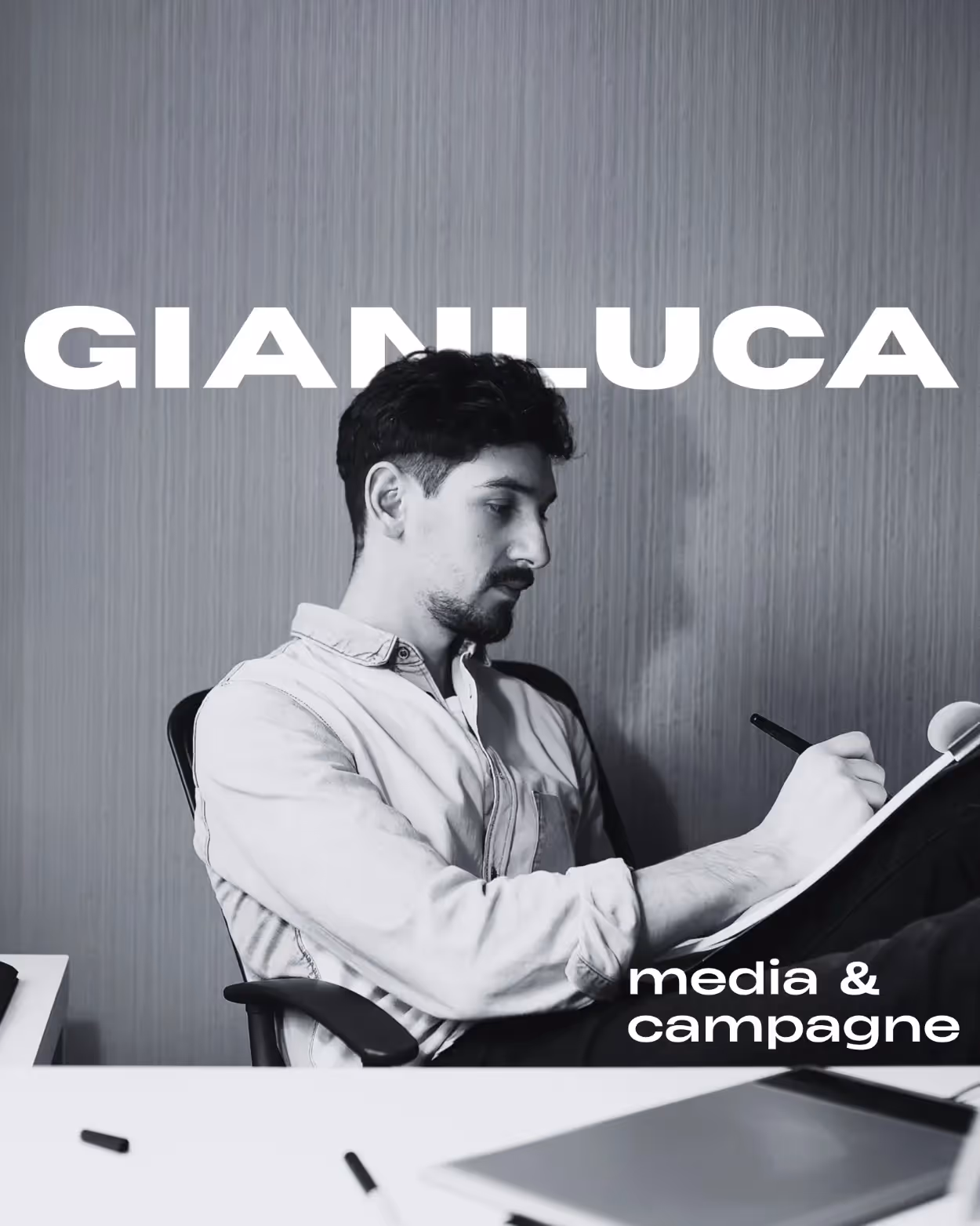 Uomo seduto alla scrivania che scrive su un blocco note, con sfondo a parete semplice e testo 'GIANLUCA' e 'media & campagne'.