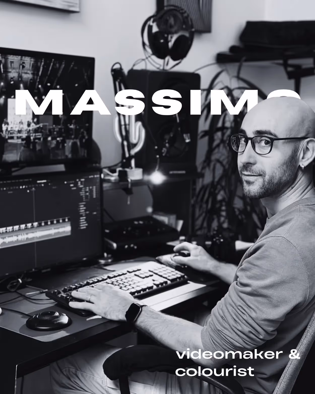 Uomo calvo con occhiali seduto a una scrivania mentre lavora al montaggio video su un computer con due monitor.