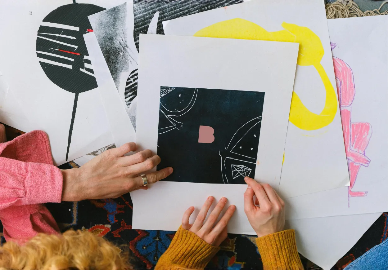 Due persone con maglioni colorati tengono stampe artistiche con forme astratte e colori vivaci su un tappeto decorato.