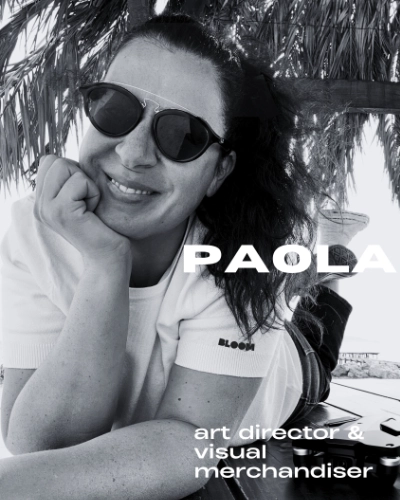 Donna sorridente con occhiali da sole, appoggiata a un supporto all'aperto sotto foglie di palma.