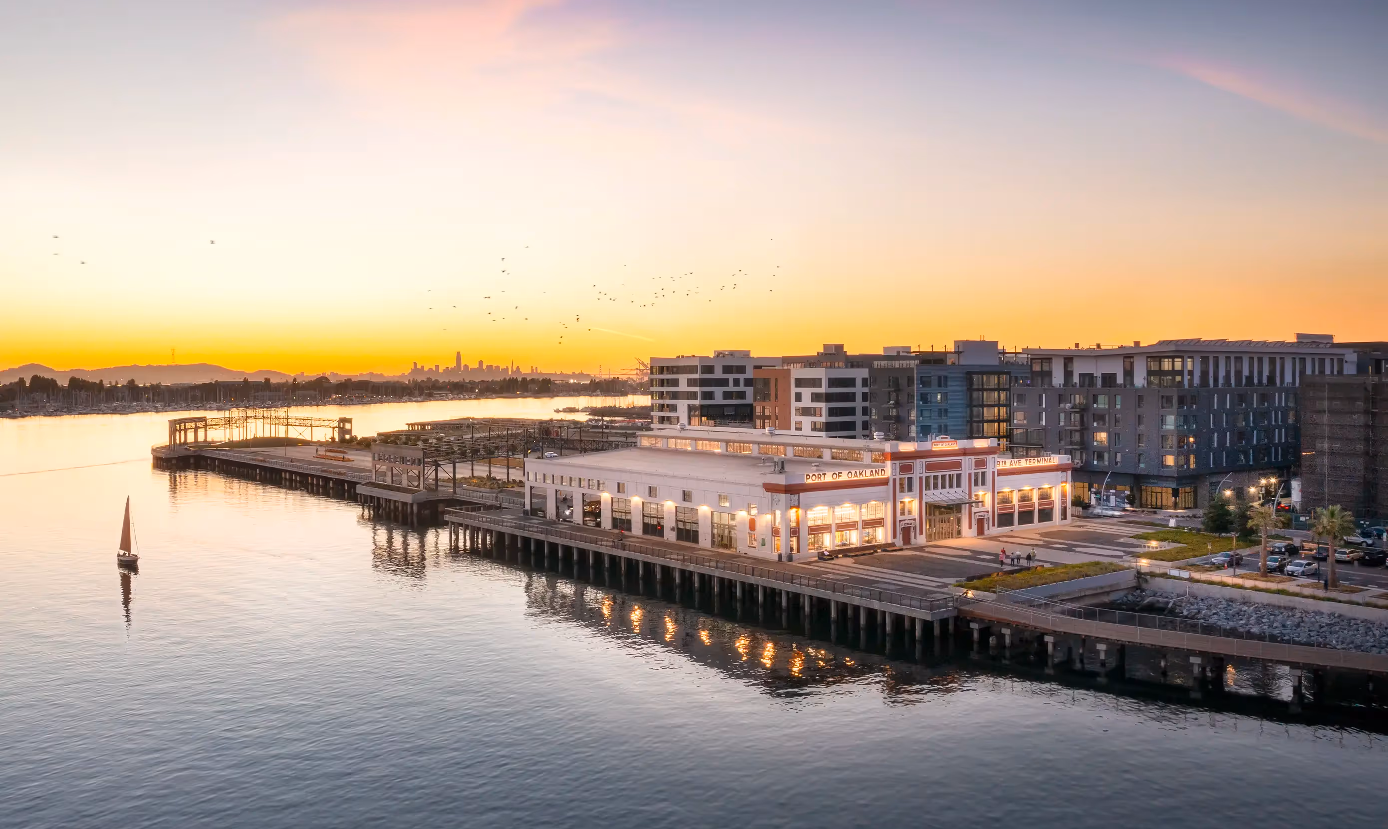 A waterfront building in Oakland with large windows sits on a pier at sunset, surrounded by modern apartment buildings and calm water; a small sailboat is nearby.