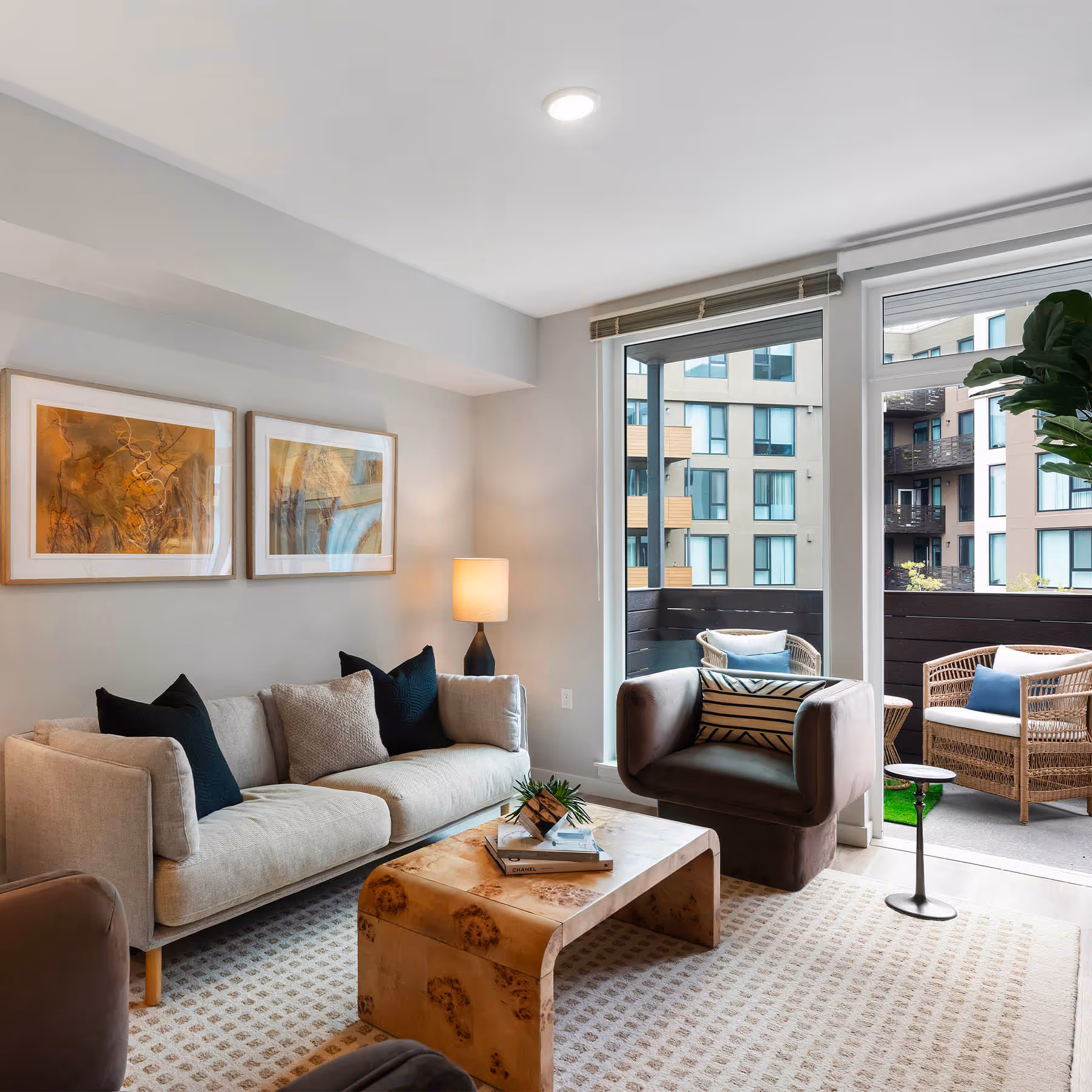 Modern living room at Caspian Residences with neutral-colored furniture, two framed artworks on the wall, two table lamps, and large windows leading to a balcony with outdoor seating.
