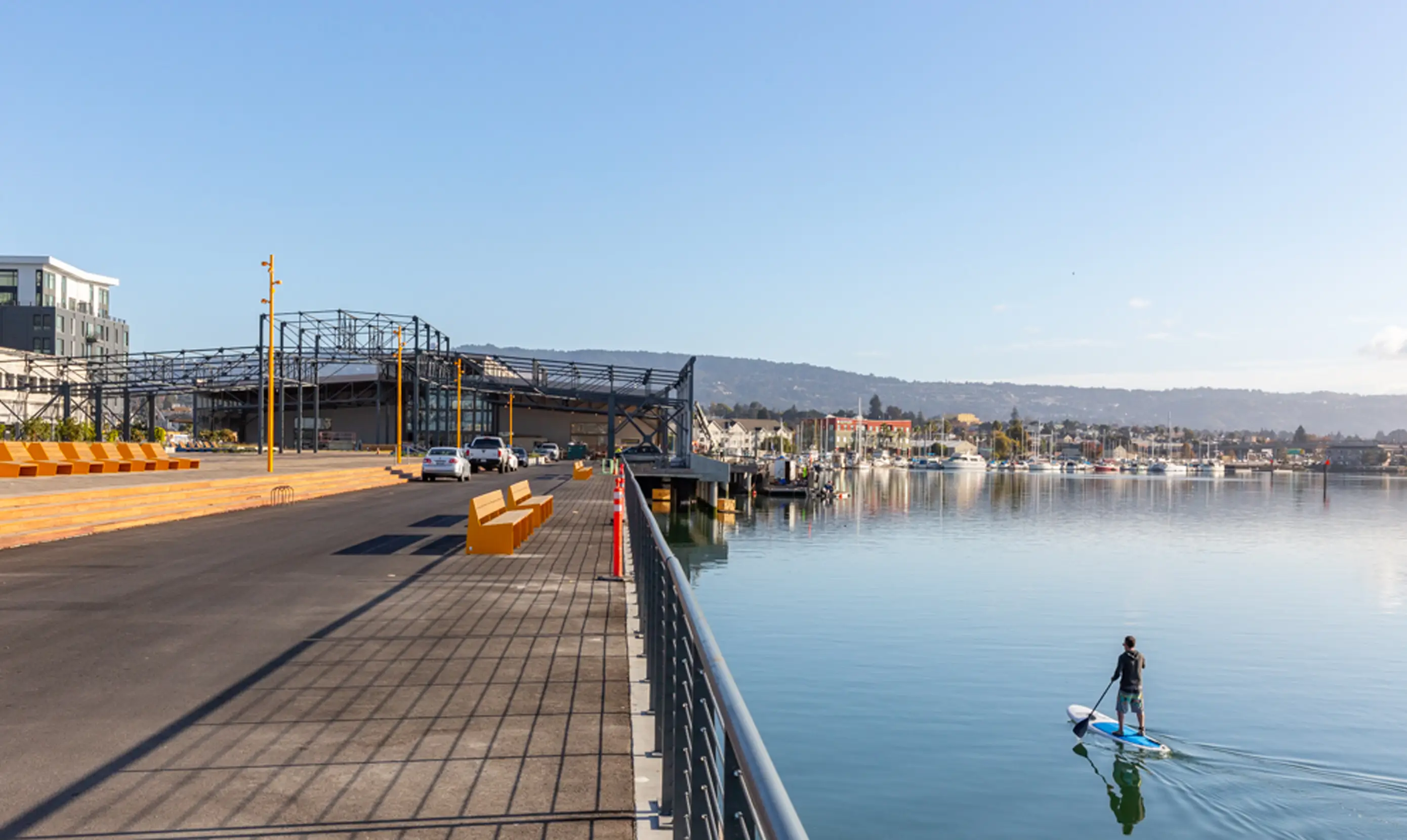 A person paddleboards on calm water near a waterfront with modern buildings, an empty promenade, and clear blue sky.