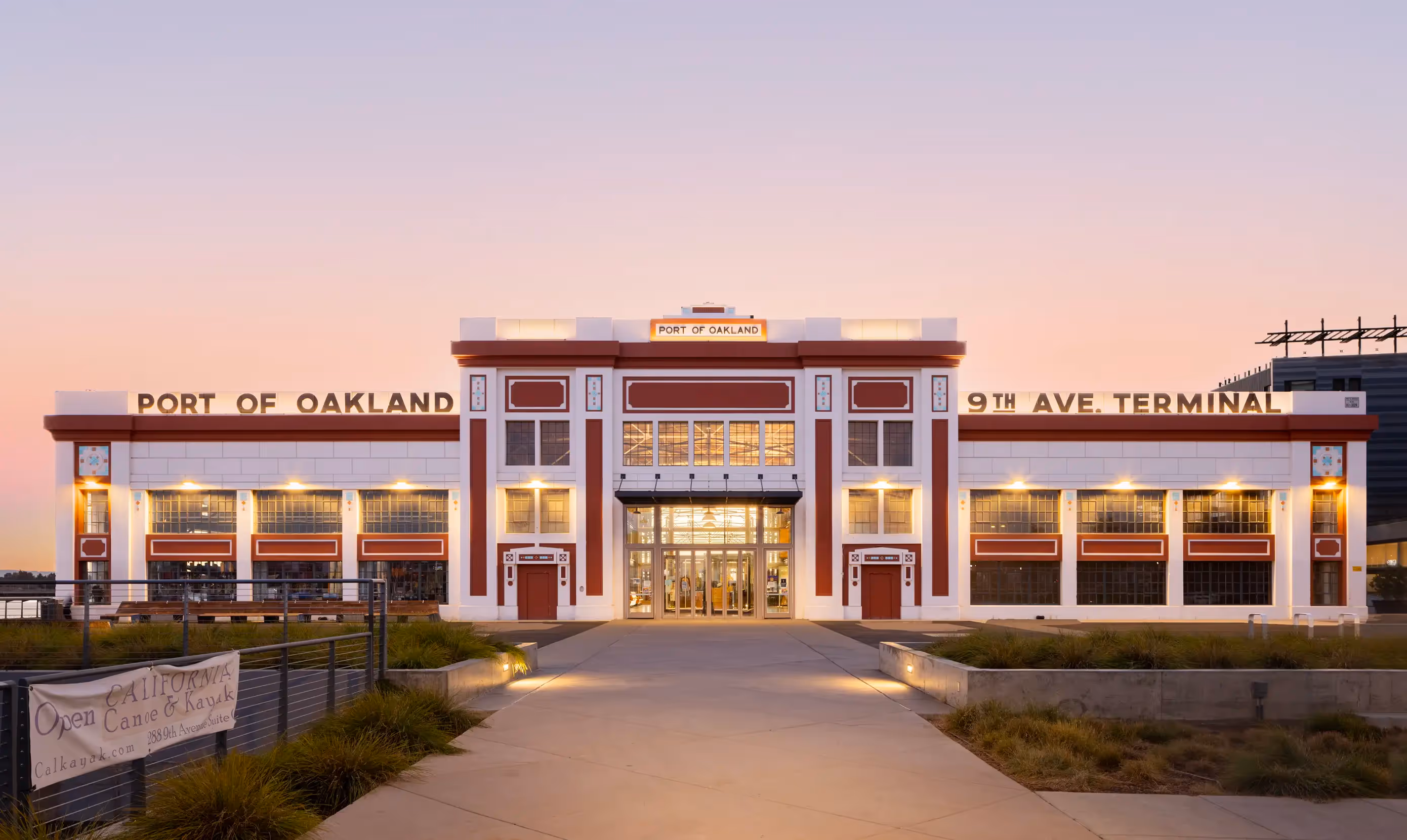 The Port of Oakland 9th Ave Terminal building at dusk, with lights on inside and a clear, pastel sky in the background.