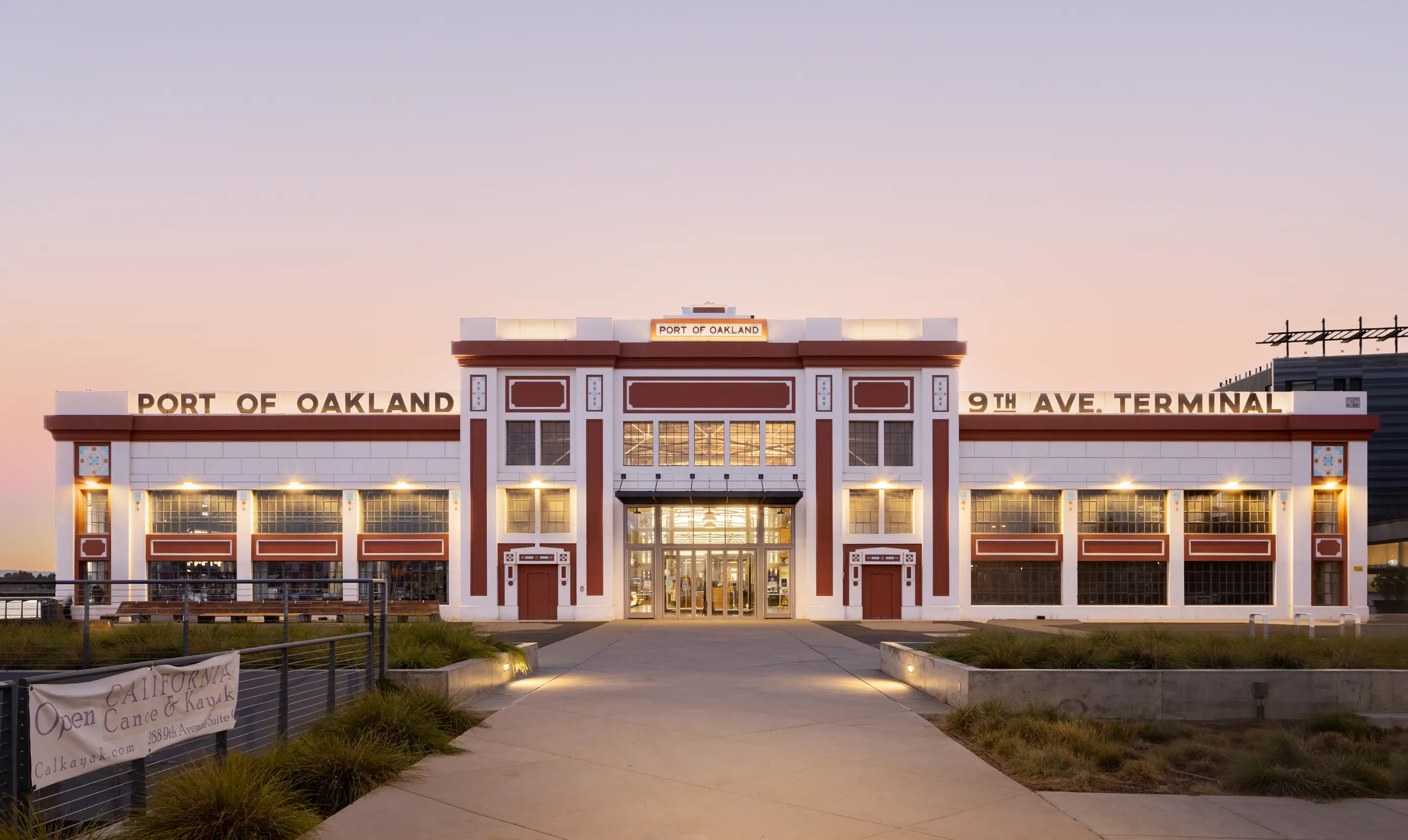 The Port of Oakland 9th Ave Terminal building at dusk, with lights on inside and a clear, pastel sky in the background.