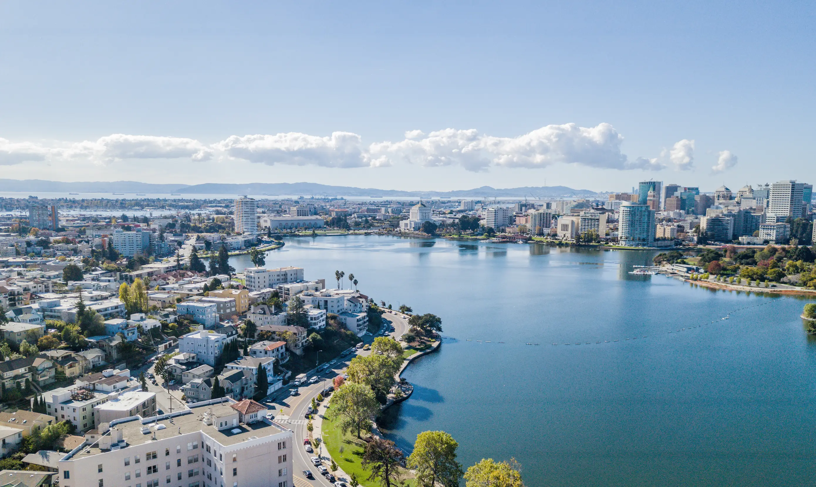 Aerial view of the Oakland skyline by a lake with buildings, trees, and mountains in the background under a clear blue sky.