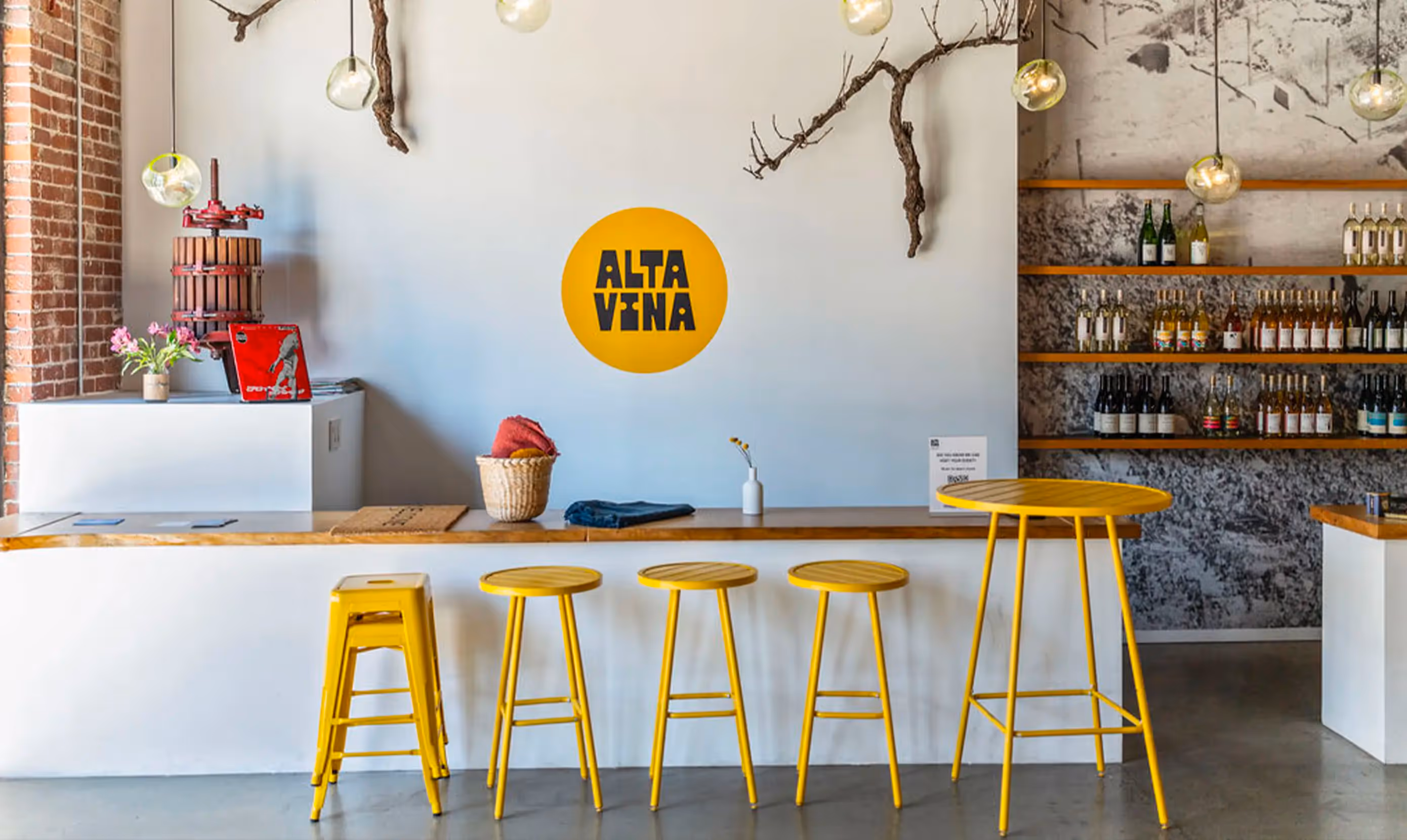  A modern bar interior featuring a white wall with a yellow circular logo that says "ALTA VINA," a wood counter, yellow stools, hanging light fixtures, a plant with flowers, and a shelf displaying various bottles of wine.