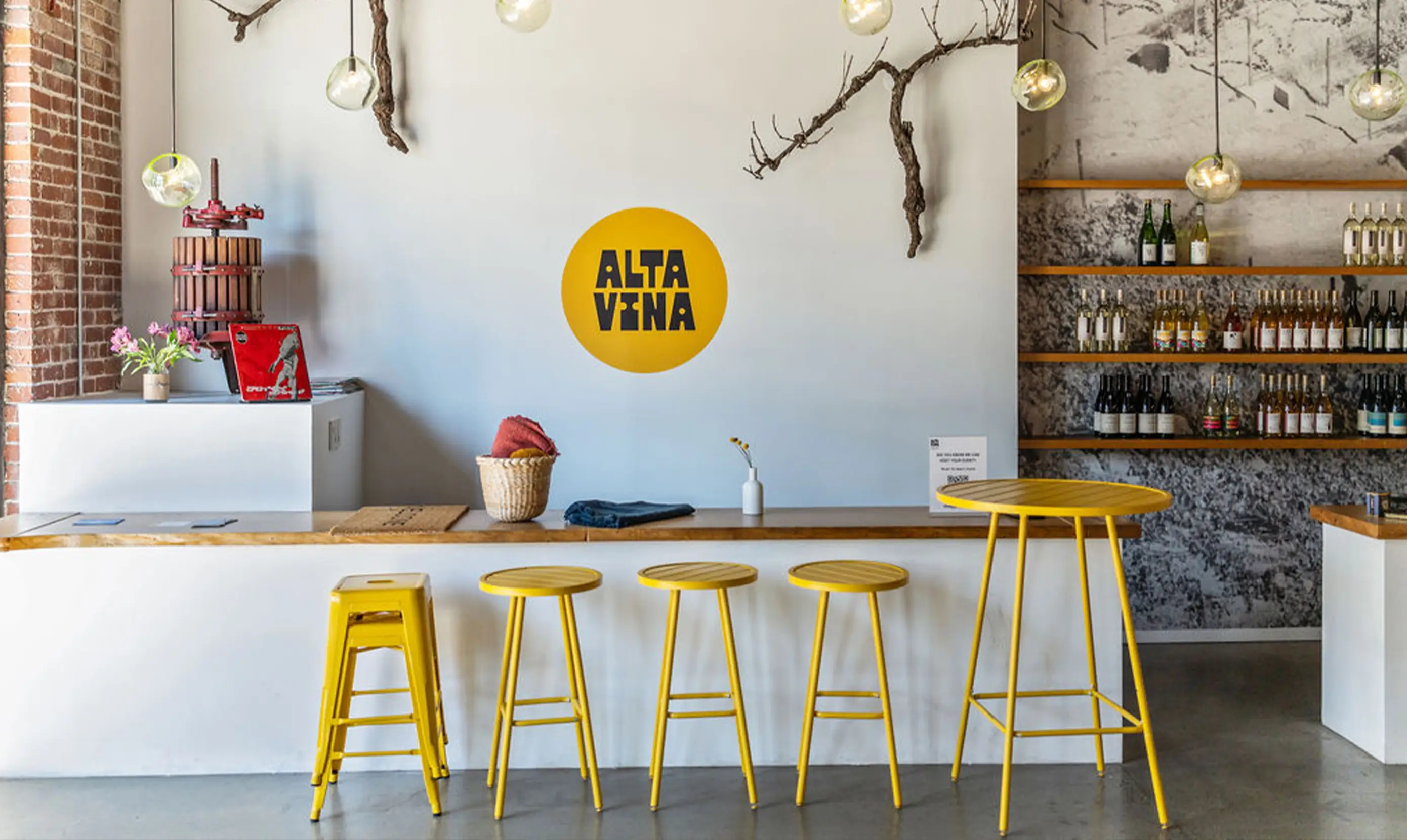  A modern bar interior featuring a white wall with a yellow circular logo that says "ALTA VINA," a wood counter, yellow stools, hanging light fixtures, a plant with flowers, and a shelf displaying various bottles of wine.