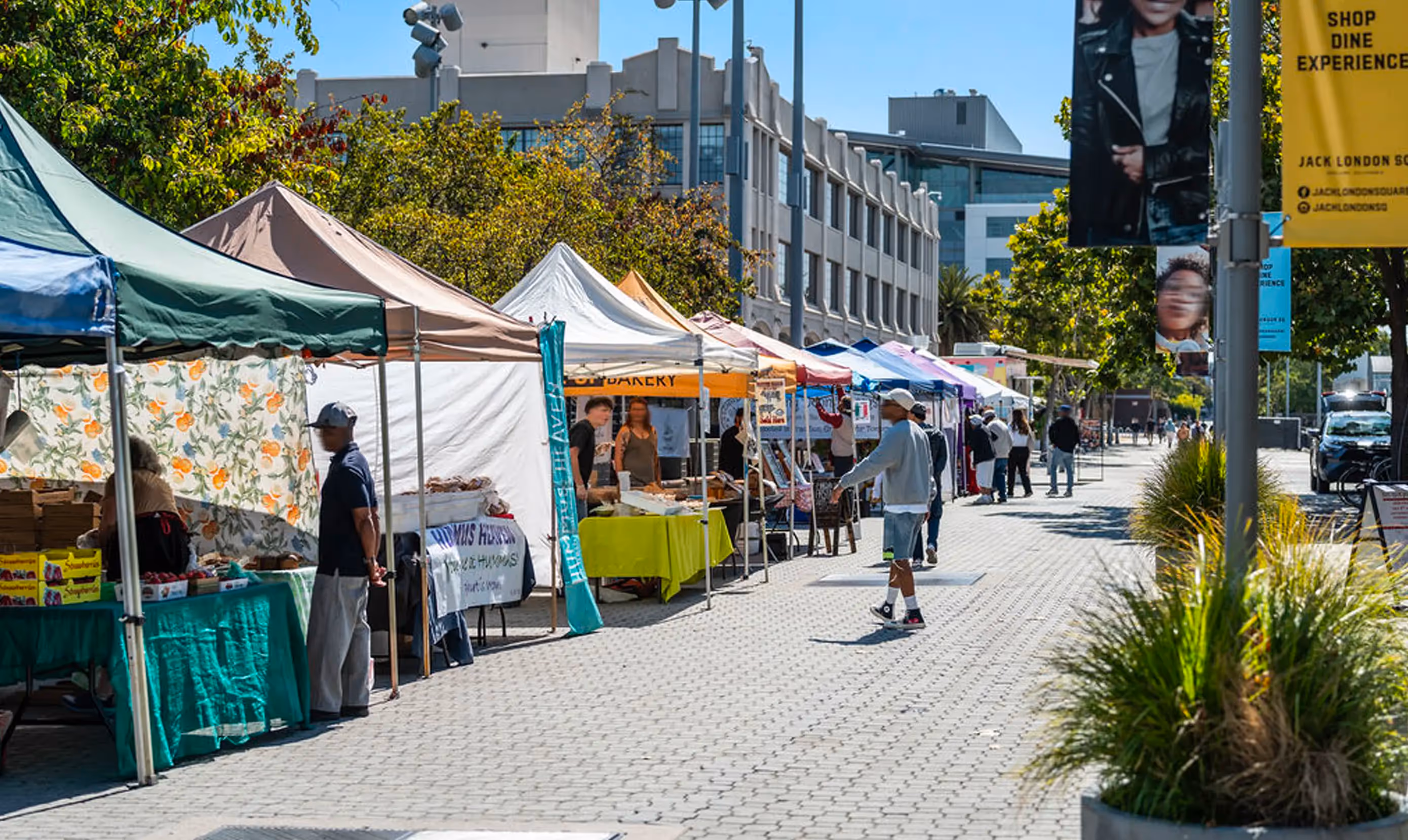  A vibrant outdoor farmers market scene with colorful tents lining a walkway, featuring vendors and shoppers on a sunny day.