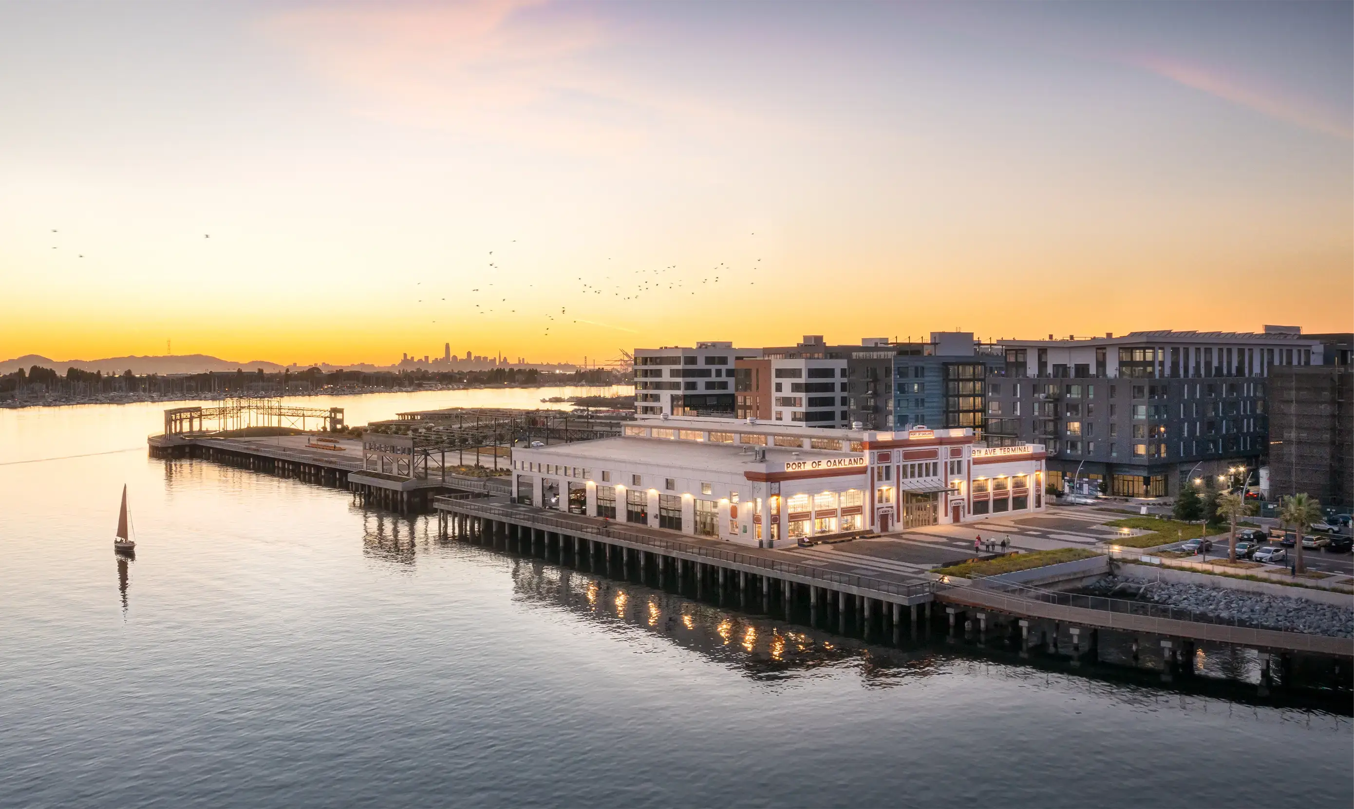 A sunset view of the Port of Oakland, featuring a sailboat in the foreground and modern buildings along the waterfront.