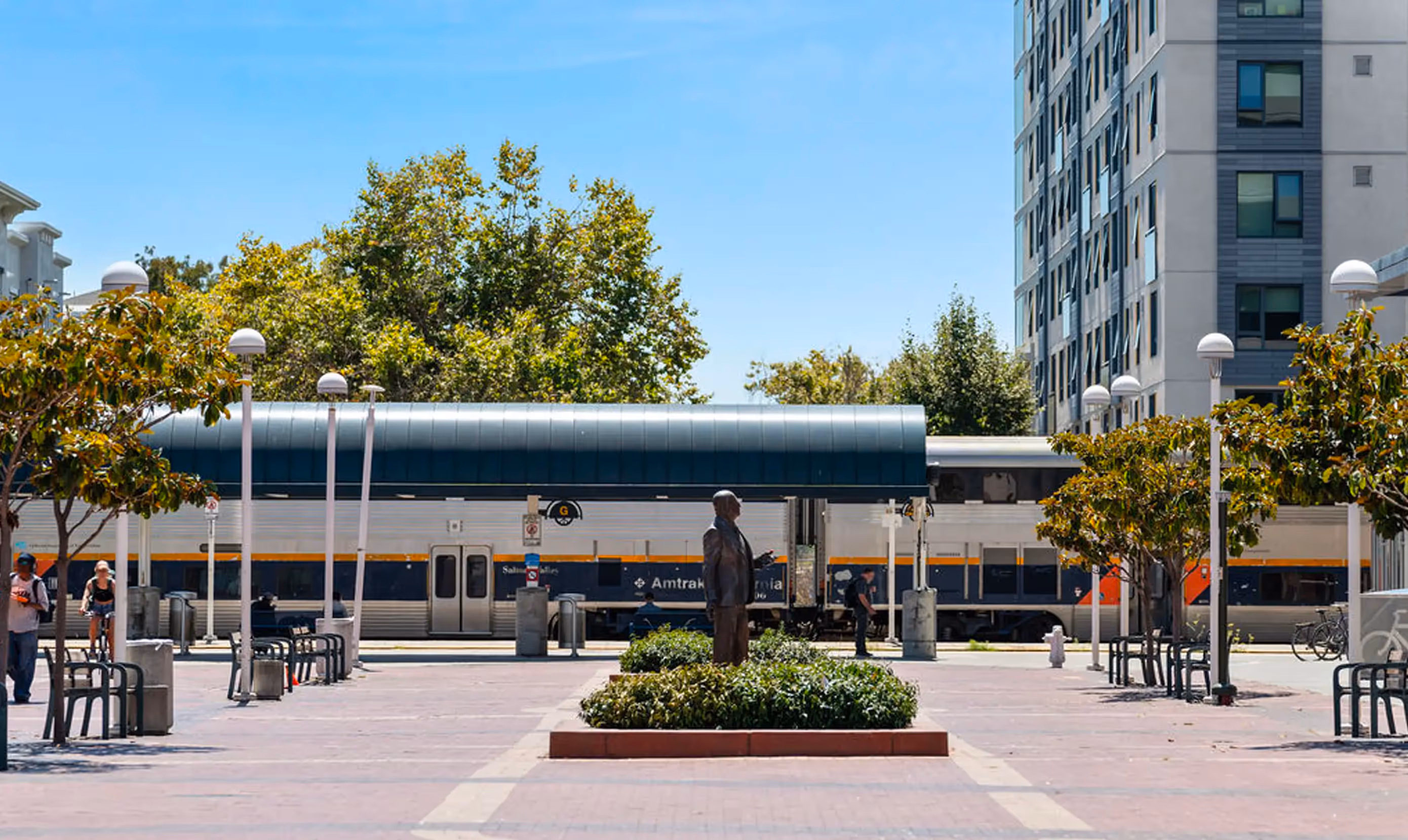 A plaza with a statue in the foreground, surrounded by trees and benches, with an Amtrak train in the background and a modern building to the right.