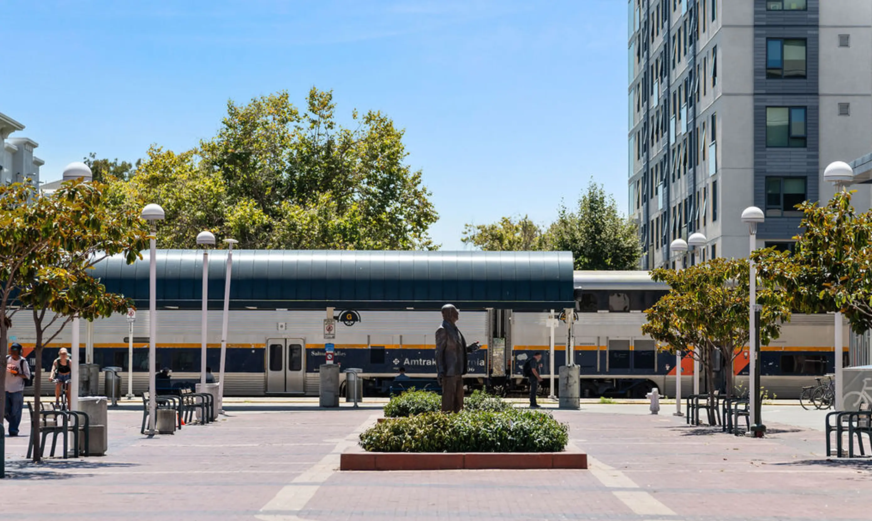 A plaza with a statue in the foreground, surrounded by trees and benches, with an Amtrak train in the background and a modern building to the right.