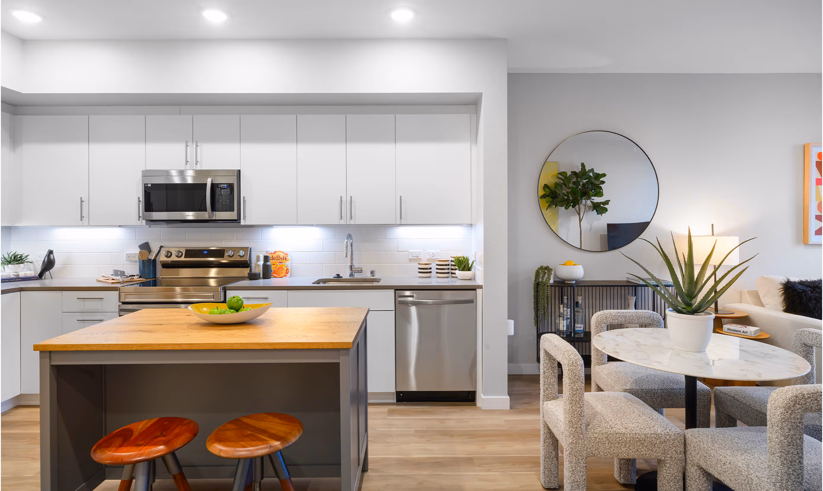 Contemporary open-concept kitchen and dining area featuring sleek white cabinetry with silver handles, stainless steel appliances including a built-in microwave, oven, and dishwasher, and a modern subway tile backsplash. A central kitchen island with a butcher-block countertop and two round wooden stools serves as both prep space and casual seating. A decorative fruit bowl with green apples and limes sits on the island, adding a pop of color. To the right, a small round marble dining table with textured boucle chairs and a potted aloe plant complements the space. The adjoining living area includes a large wall mirror, console table with decor, and part of a beige sectional sofa with accent pillows. Warm wood flooring and recessed lighting enhance the bright, clean, and modern apartment design.