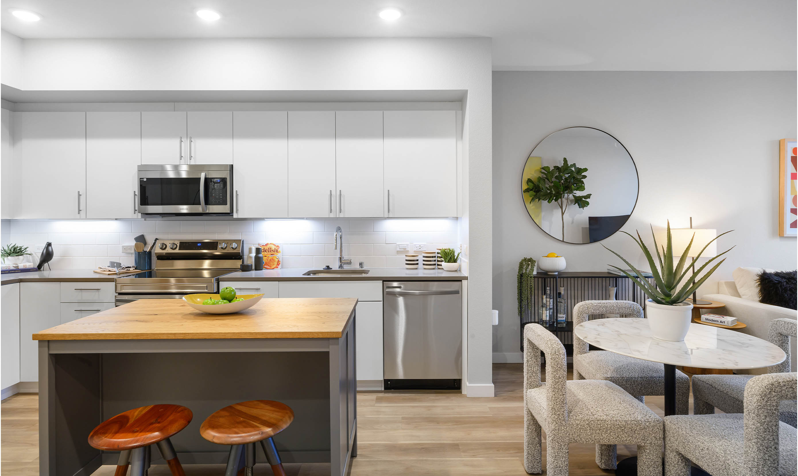 Contemporary open-concept kitchen and dining area featuring sleek white cabinetry with silver handles, stainless steel appliances including a built-in microwave, oven, and dishwasher, and a modern subway tile backsplash. A central kitchen island with a butcher-block countertop and two round wooden stools serves as both prep space and casual seating. A decorative fruit bowl with green apples and limes sits on the island, adding a pop of color. To the right, a small round marble dining table with textured boucle chairs and a potted aloe plant complements the space. The adjoining living area includes a large wall mirror, console table with decor, and part of a beige sectional sofa with accent pillows. Warm wood flooring and recessed lighting enhance the bright, clean, and modern apartment design.