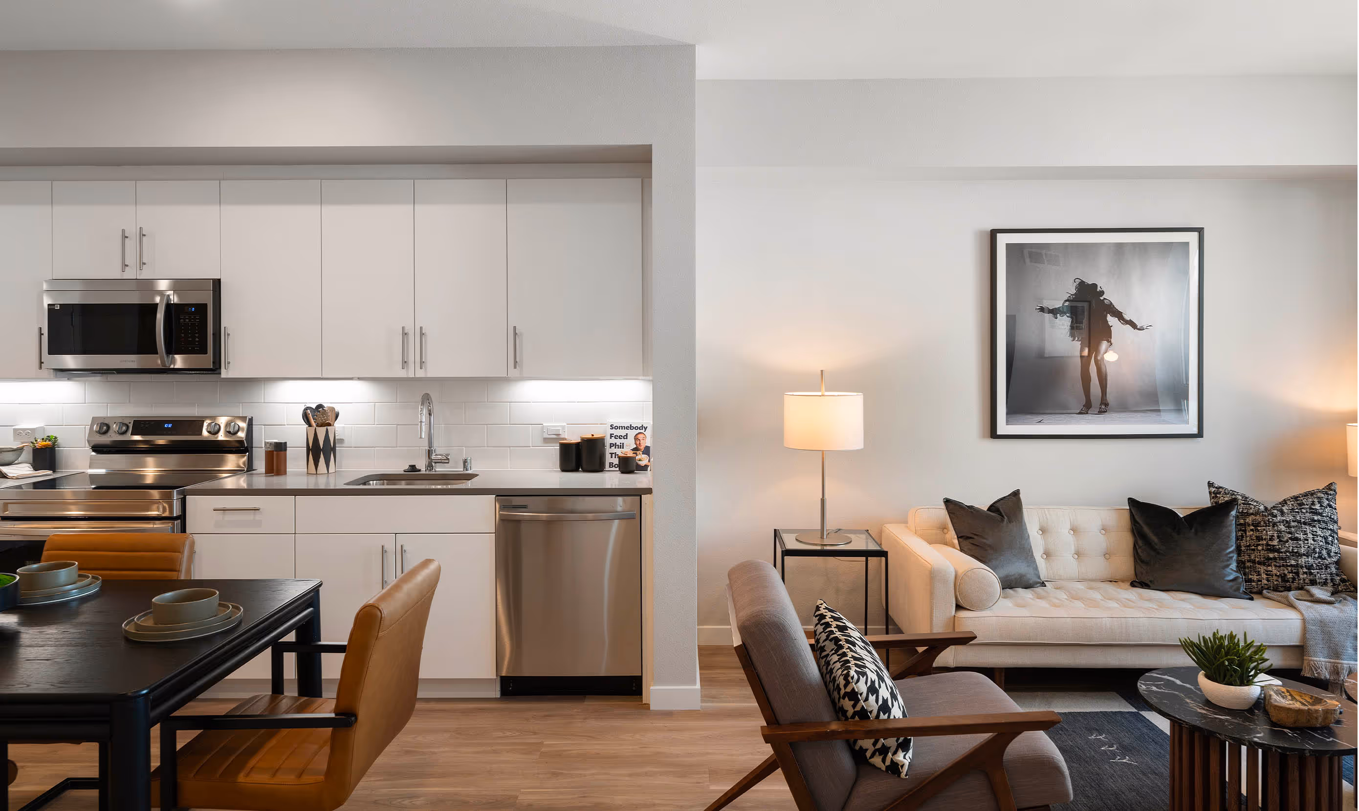 Modern open-plan kitchen and living area with wood flooring. White cabinets, stainless steel appliances, cozy sofa, chic art, and warm lighting create a welcoming feel.