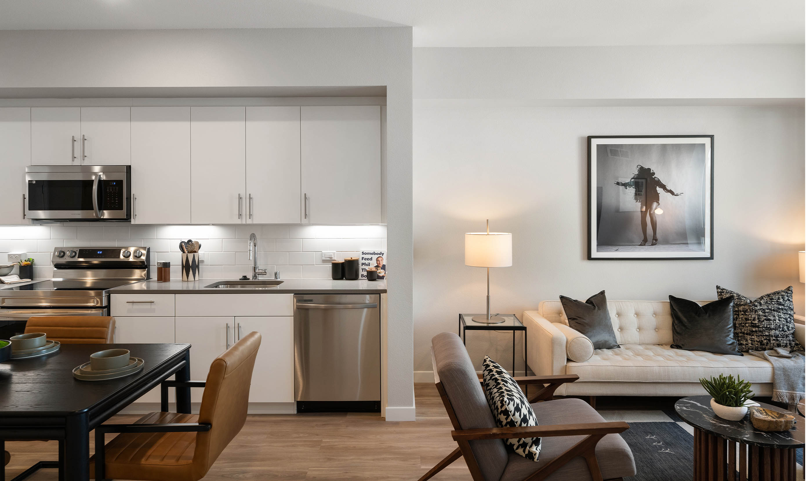 Modern open-plan kitchen and living area with wood flooring. White cabinets, stainless steel appliances, cozy sofa, chic art, and warm lighting create a welcoming feel.