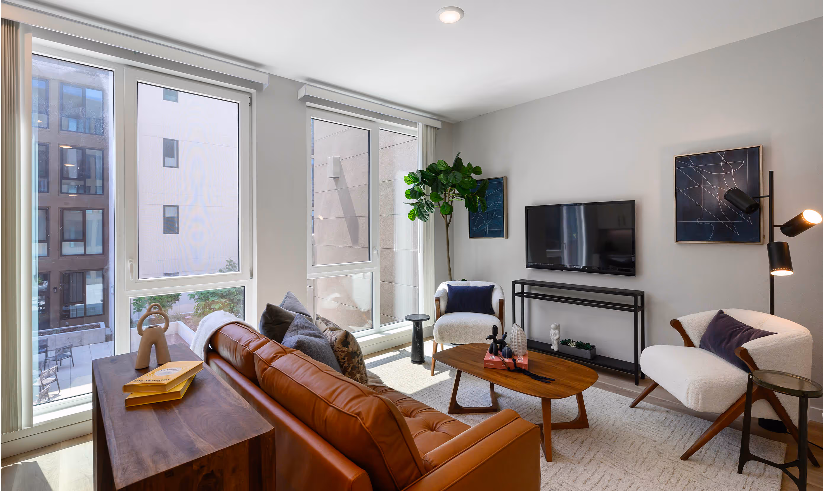 Modern living room with large windows, a brown leather sofa, two white armchairs, and a TV on a sleek stand. A tall plant adds greenery, while abstract art, a wooden coffee table, and a floor lamp create a cozy, stylish ambiance.