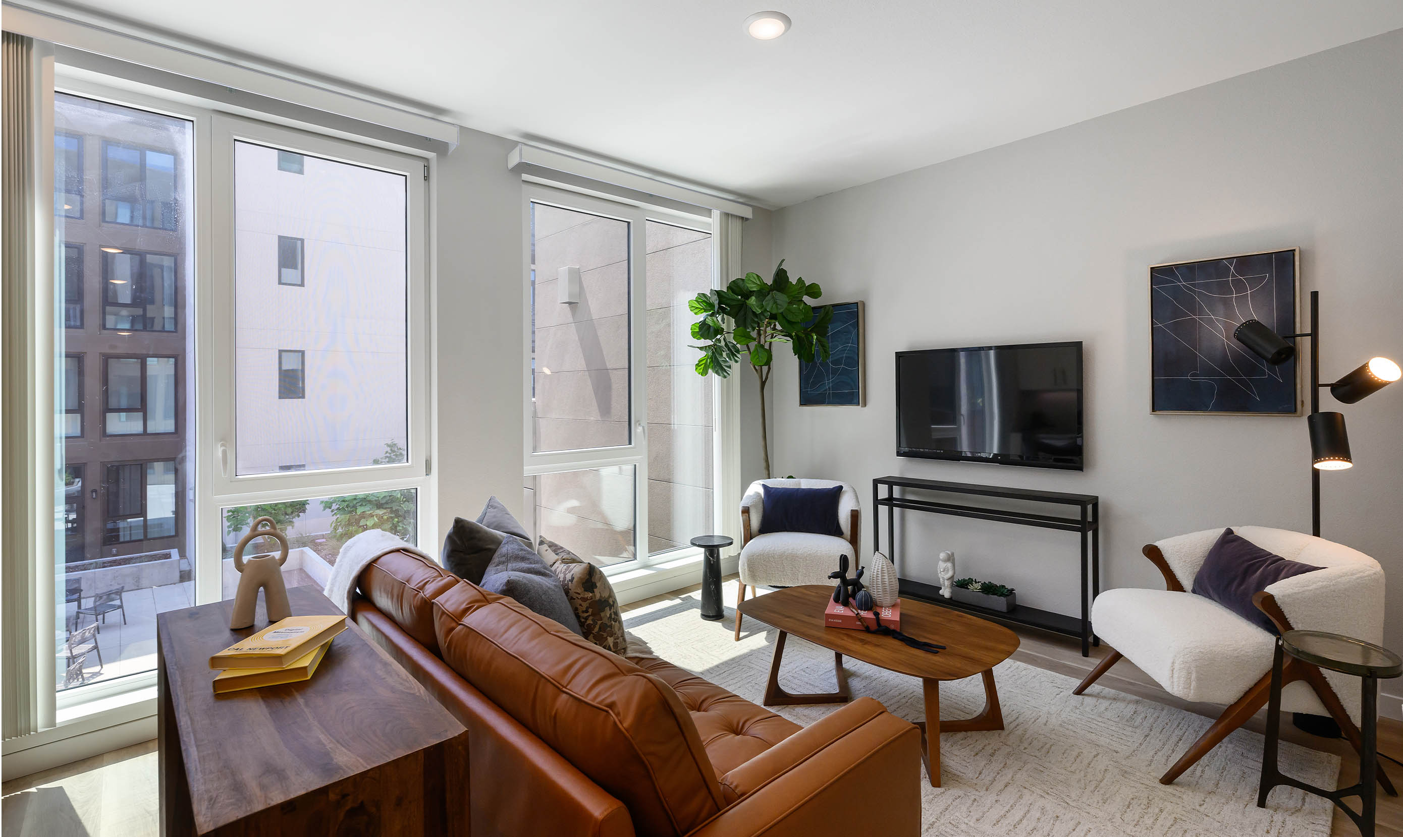 Modern living room with large windows, a brown leather sofa, two white armchairs, and a TV on a sleek stand. A tall plant adds greenery, while abstract art, a wooden coffee table, and a floor lamp create a cozy, stylish ambiance.