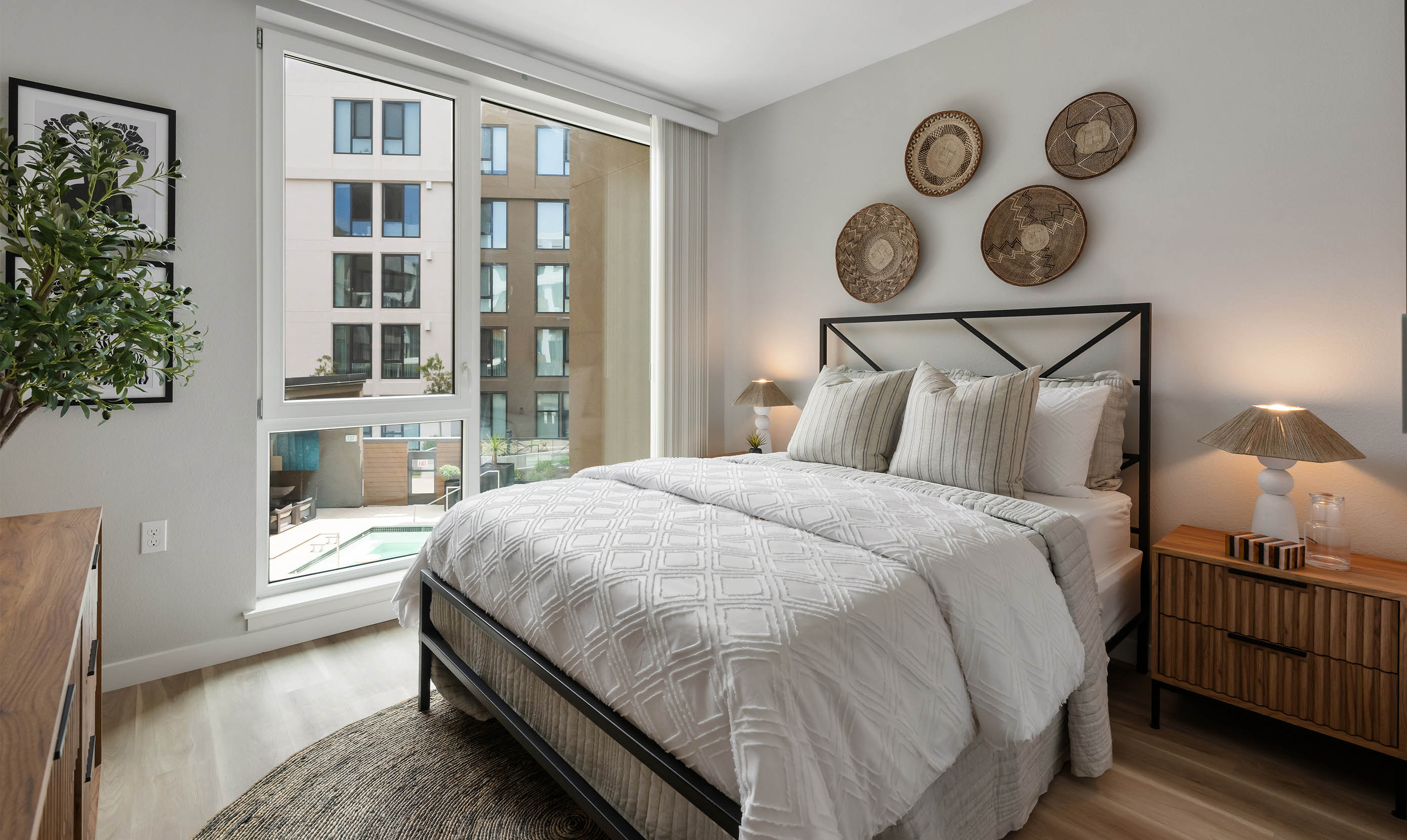 A modern bedroom features a neatly made bed with white bedding, wooden nightstands, and soft lighting. Large windows reveal a view of urban buildings.