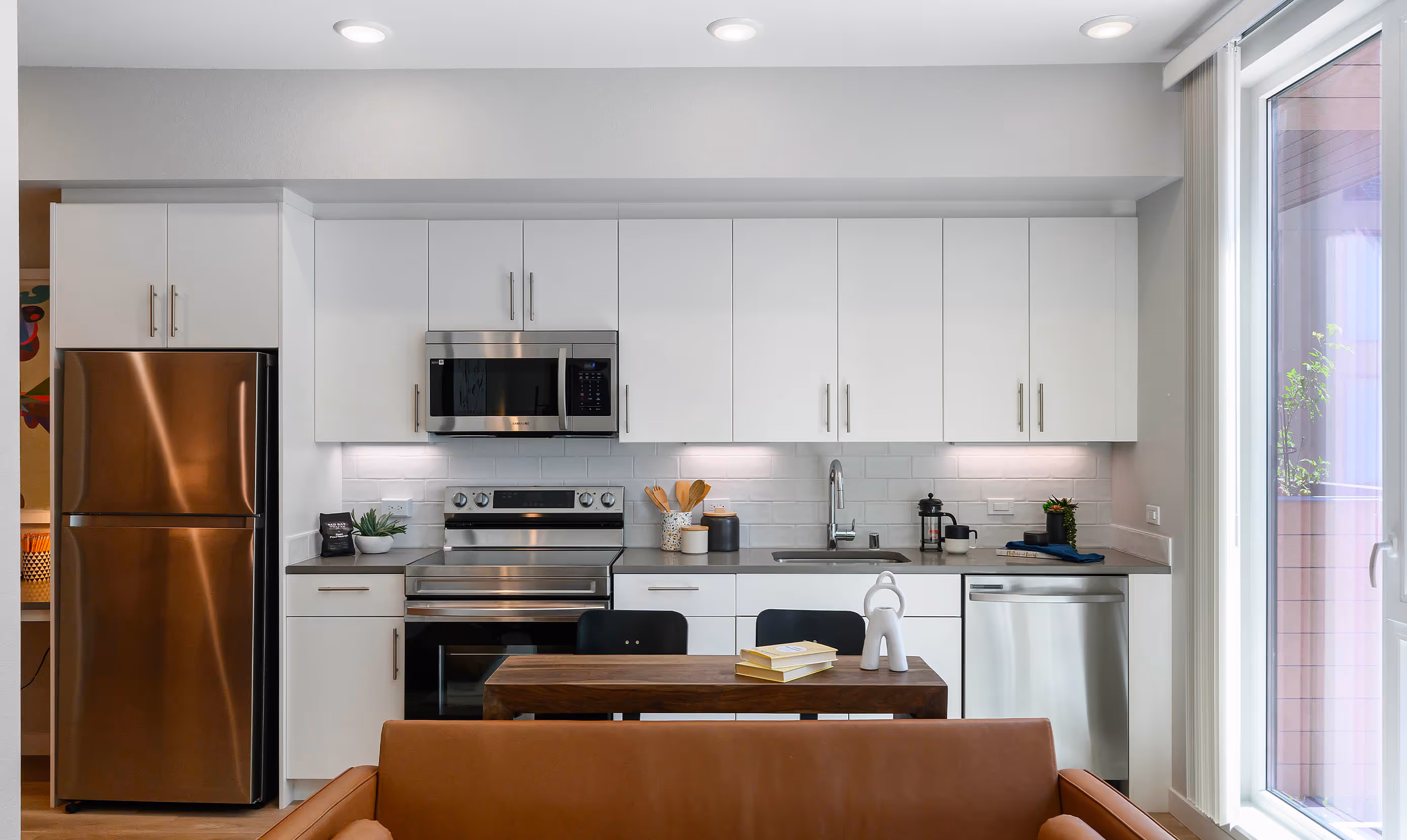Bright modern kitchen with white cabinets, stainless steel appliances, and a cozy brown leather sofa in the foreground. Calm, inviting atmosphere.