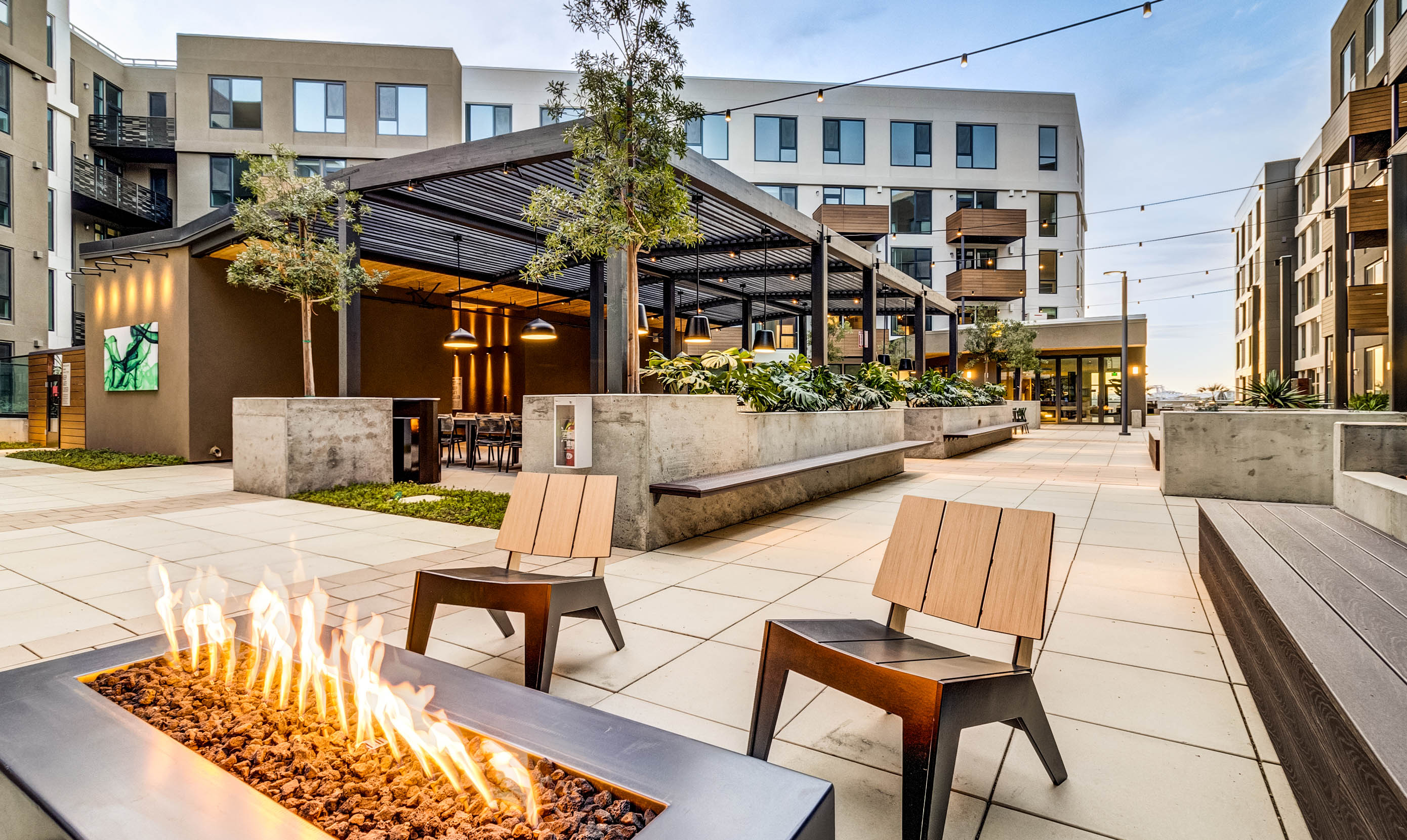 Modern apartment courtyard with a stylish fire pit, and wooden chairs. Surrounded by greenery, a covered seating area, and strings of lights.