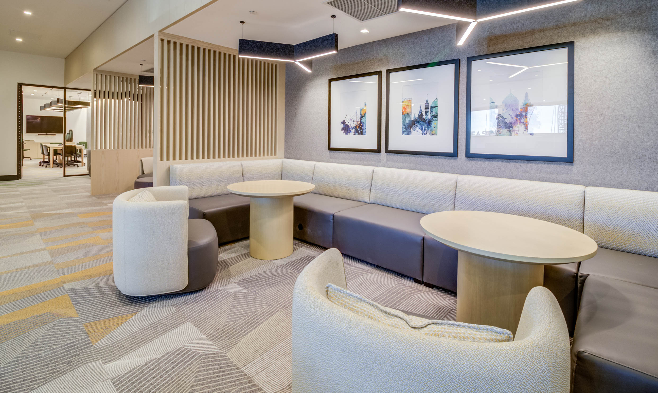 Modern office lounge with neutral tones, featuring a corner sofa, round tables, abstract art on walls, and soft lighting, creating a calm atmosphere.
