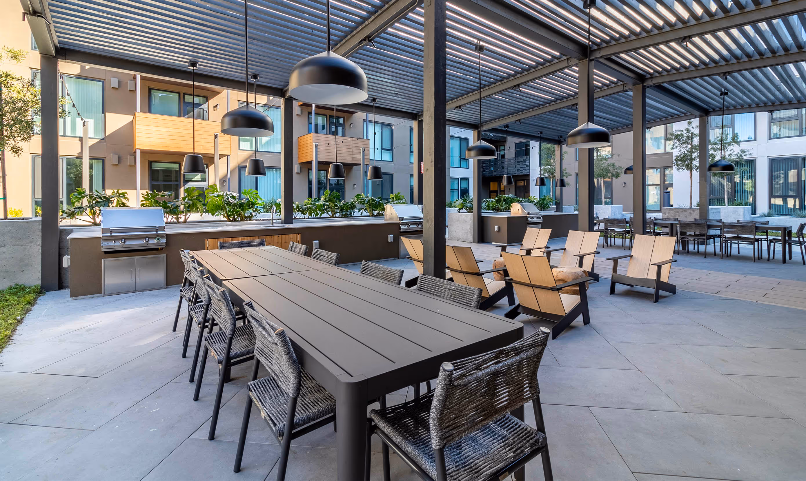 Outdoor patio with modern seating includes a long table, wicker chairs, and wooden lounge chairs under a pergola. Buildings and plants surround the space.