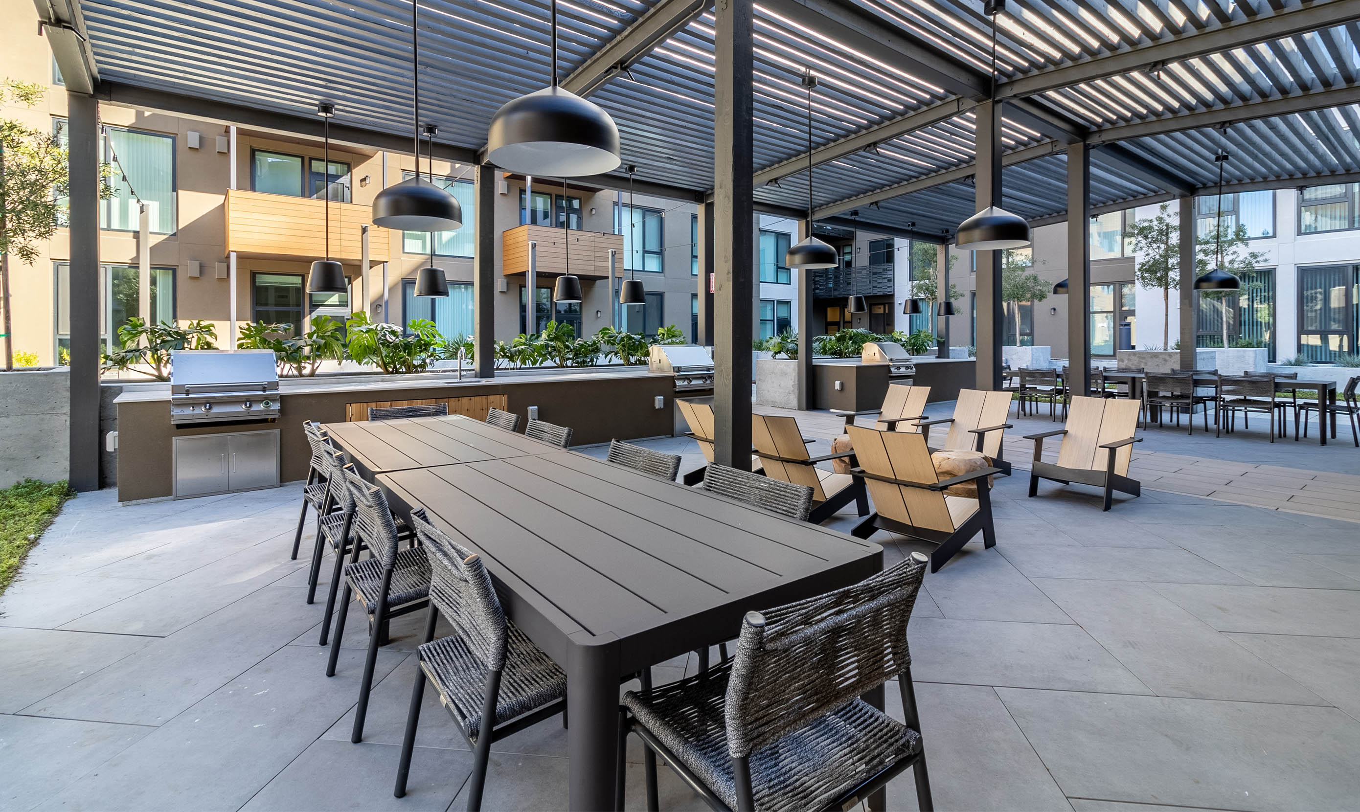 Outdoor patio with modern seating includes a long table, wicker chairs, and wooden lounge chairs under a pergola. Buildings and plants surround the space.