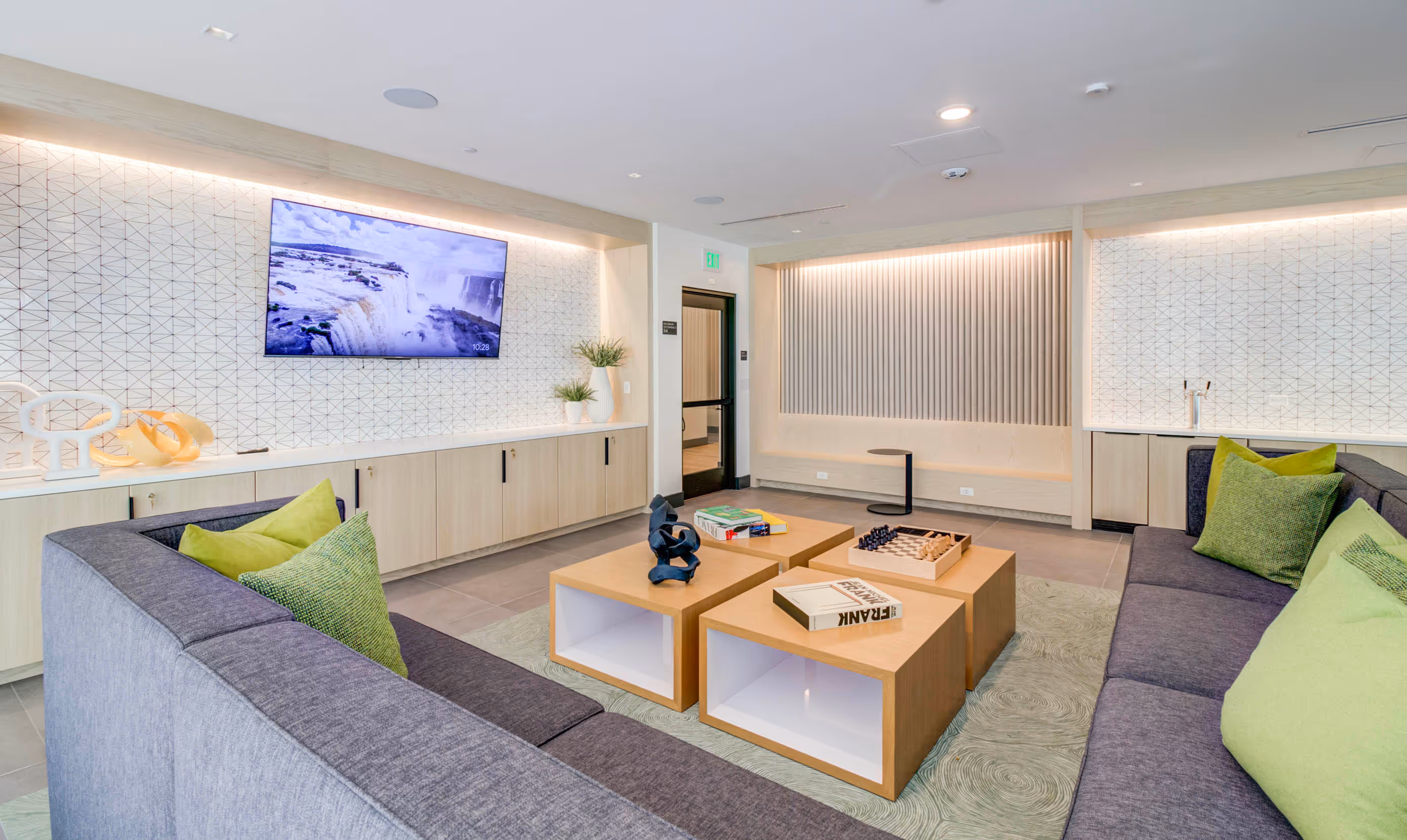 A modern lounge with a gray sectional sofa, green cushions, and wooden tables. A TV is on a patterned wall, creating a cozy, inviting atmosphere.