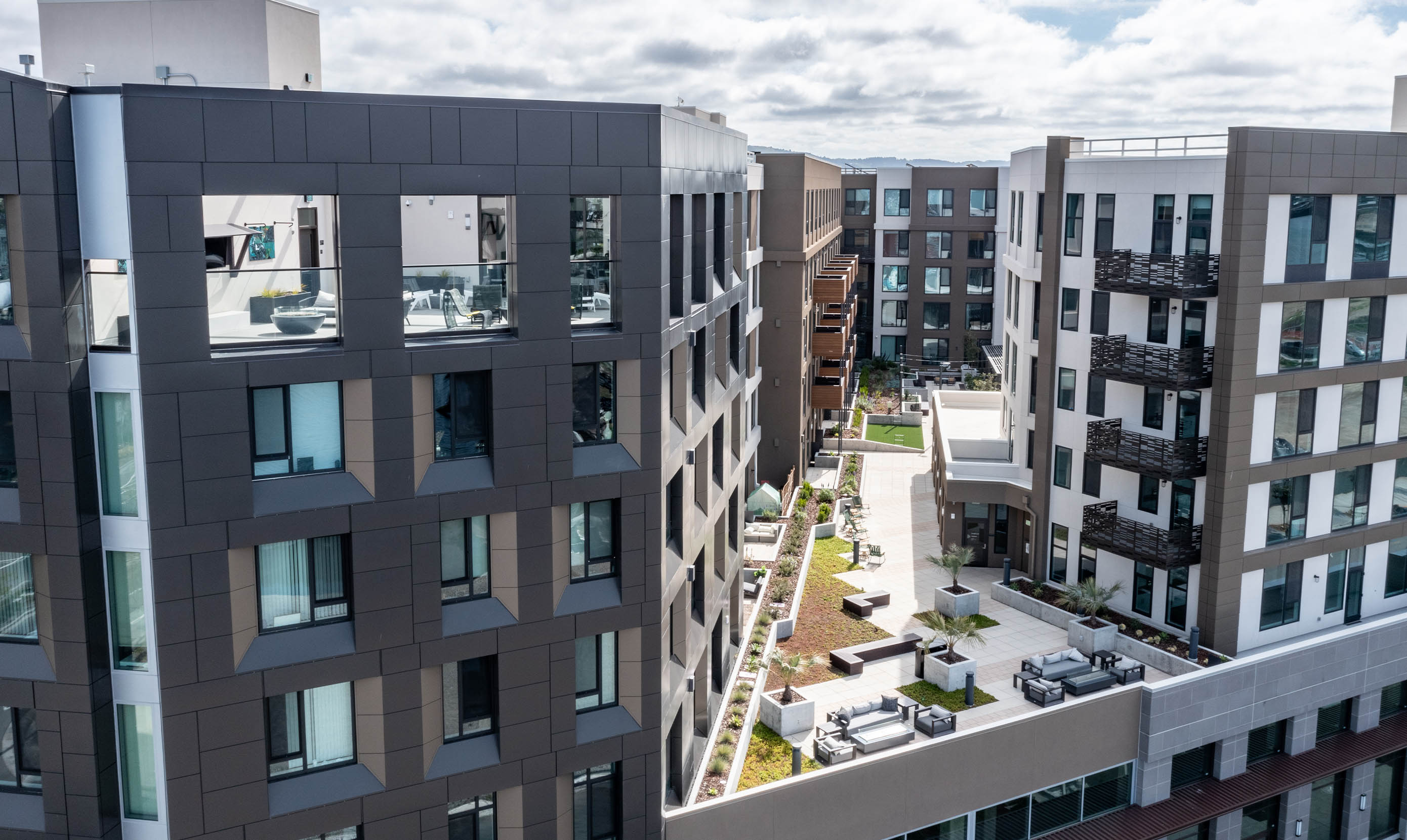 Modern apartment complex with rectangular buildings featuring balconies and large windows. Central courtyard with greenery and seating areas, under a partly cloudy sky.