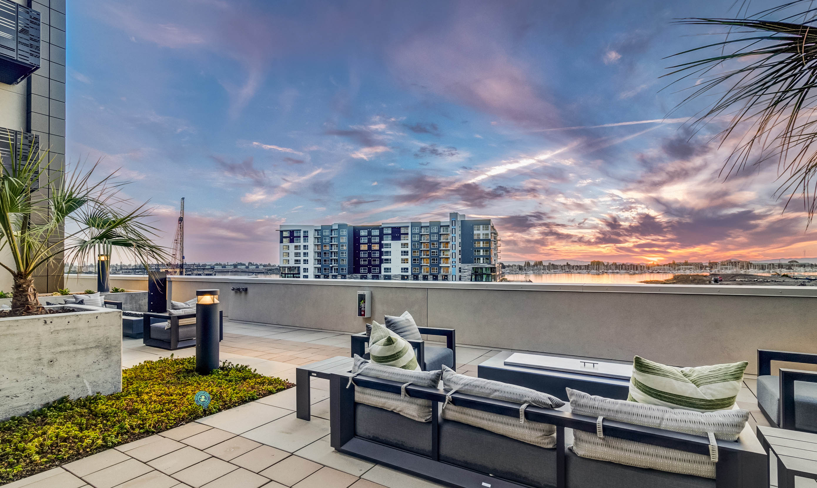 Rooftop terrace with stylish lounge furniture overlooks a scenic sunset. Vibrant sky, modern cityscape, and water view create a serene ambiance.