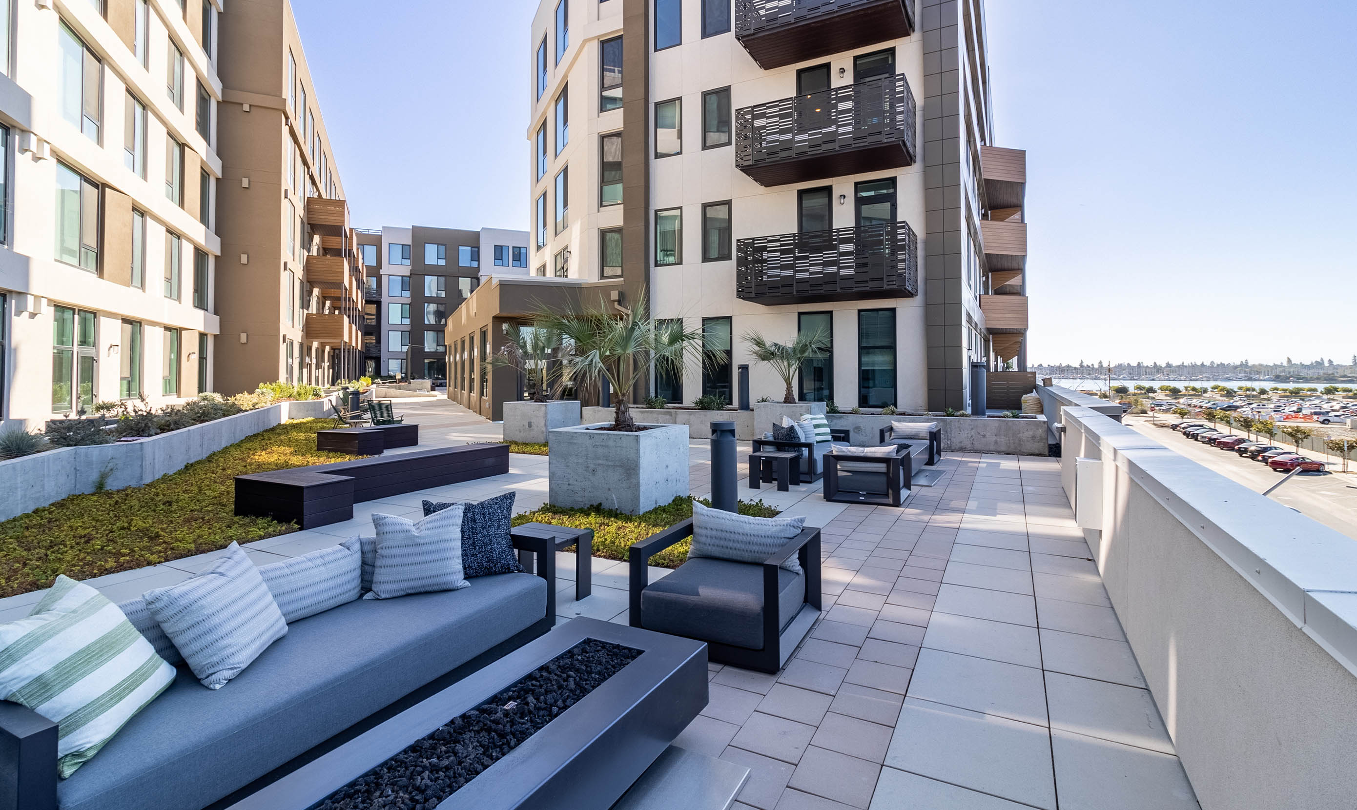 Modern rooftop terrace with gray cushioned seating, fire pits, and planters. Surrounding buildings add an urban vibe under a clear blue sky.
