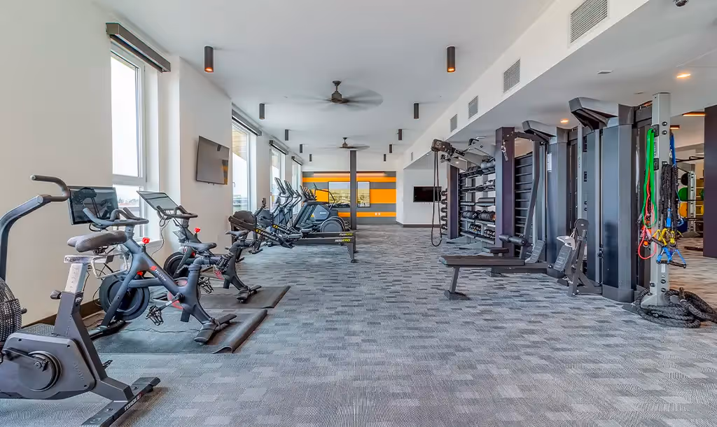 A modern gym with exercise bikes, treadmills, and strength training equipment in a spacious, well-lit room. Large windows and sleek decor create an inviting atmosphere.