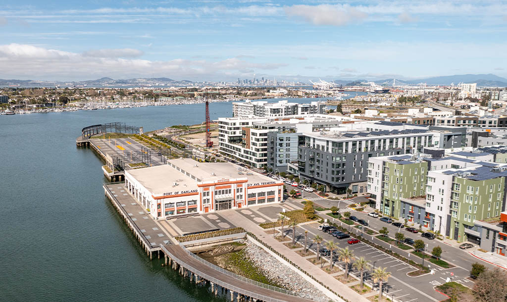 Aerial view of Oakland waterfront featuring modern buildings, a long pier, and a calm bay. The scene conveys a vibrant, urban atmosphere under a clear blue sky.