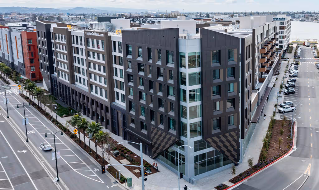 Aerial view of a modern, multi-story apartment complex on a street corner, with sleek design, palm trees lining the road, and a calm urban atmosphere.