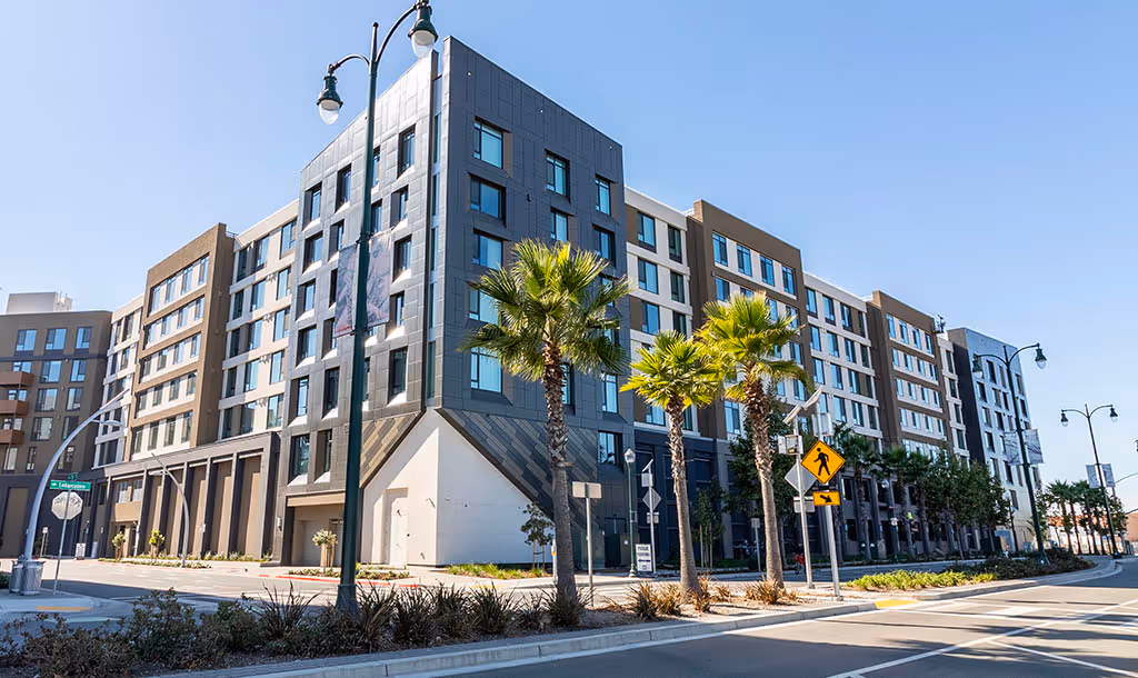A modern, multi-story residential building with large windows and a sleek design, surrounded by palm trees and streetlights under a clear blue sky.