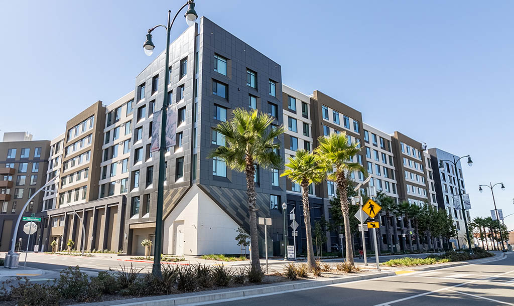 A modern, multi-story residential building with large windows and a sleek design, surrounded by palm trees and streetlights under a clear blue sky.